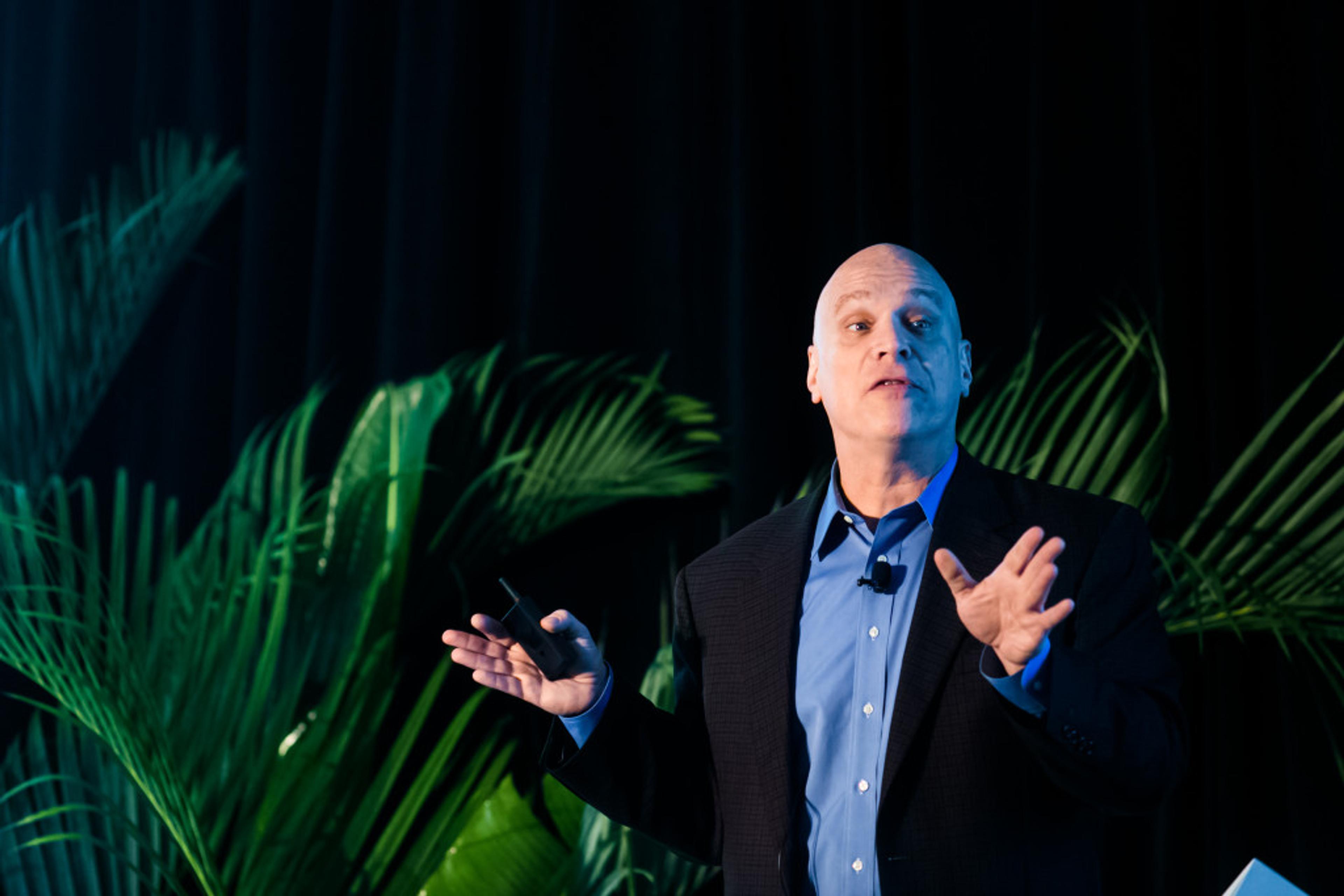 A speaker gestures while presenting on stage, with green palm leaves in the background. The speaker is wearing a dark blazer and a blue shirt.