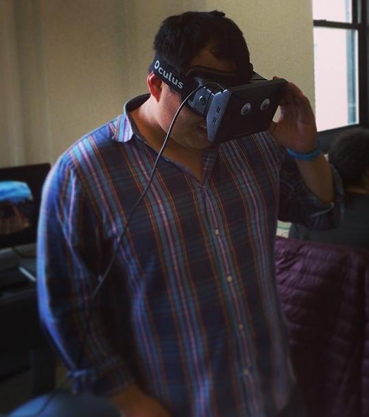 A person wearing an Oculus virtual reality headset stands indoors, appearing focused on the experience. The background shows a computer setup.