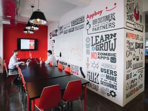 A modern conference room with a long table and red chairs, featuring a wall covered in motivational phrases and graphics. Two people are seated at the table, working on laptops.
