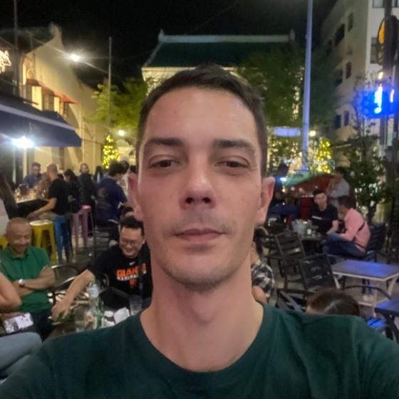 A man takes a selfie in a lively outdoor setting with people seated at tables in the background. The scene is illuminated by colorful lights at night.