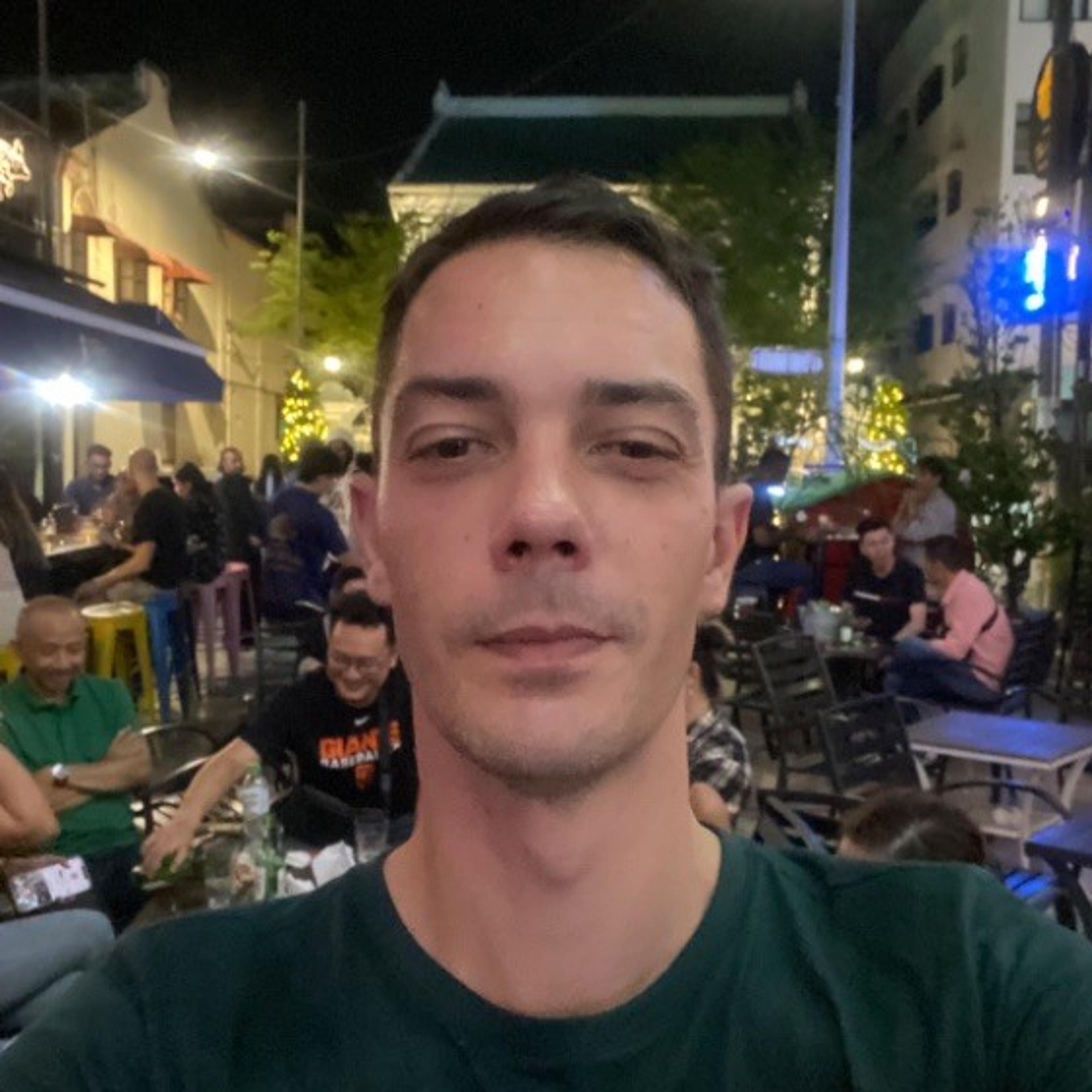 A man takes a selfie in a lively outdoor setting with people seated at tables in the background. The scene is illuminated by colorful lights at night.