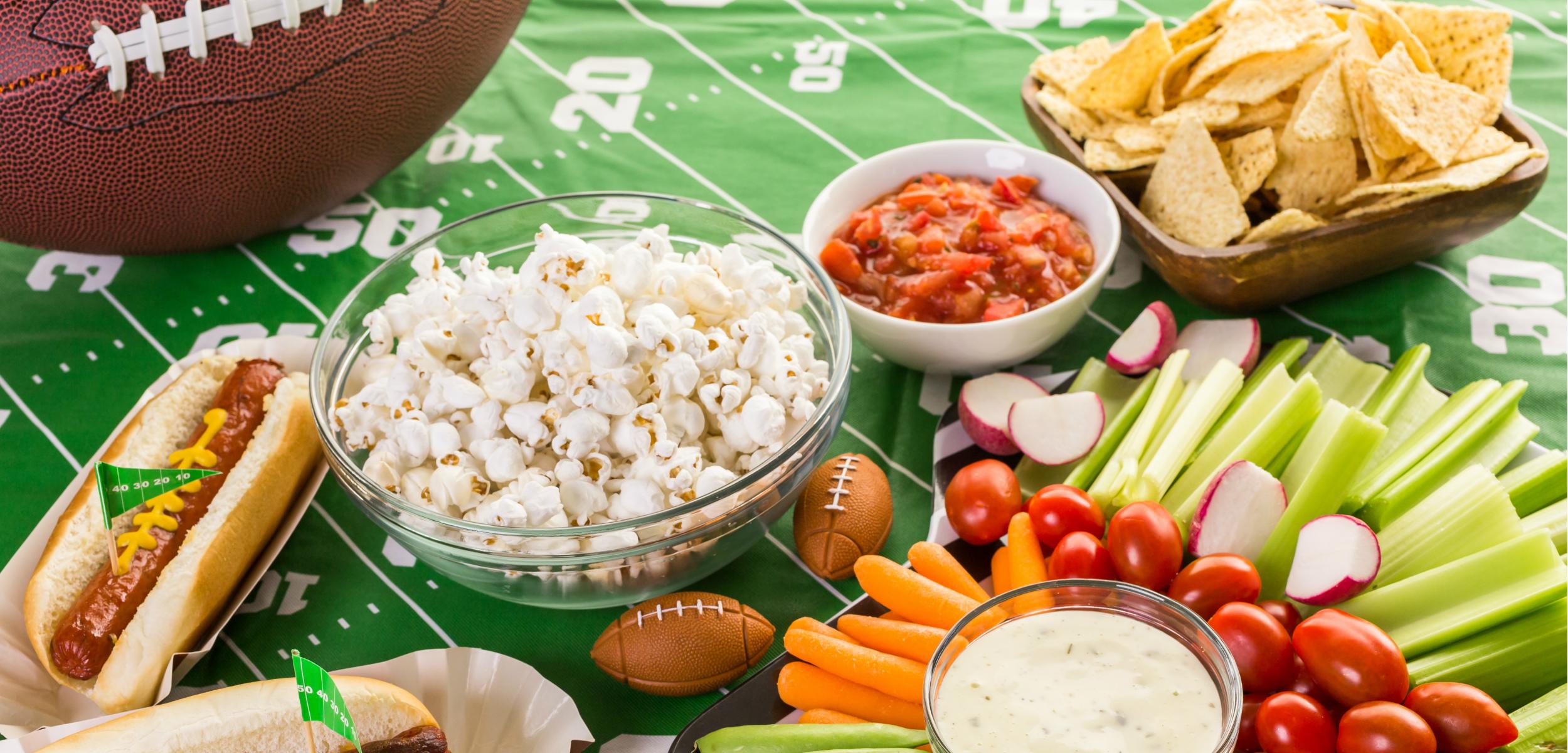 A spread of game day snacks including popcorn, hot dogs, salsa, tortilla chips, and fresh vegetables on a football-themed tablecloth. Small football decorations are scattered among the food.