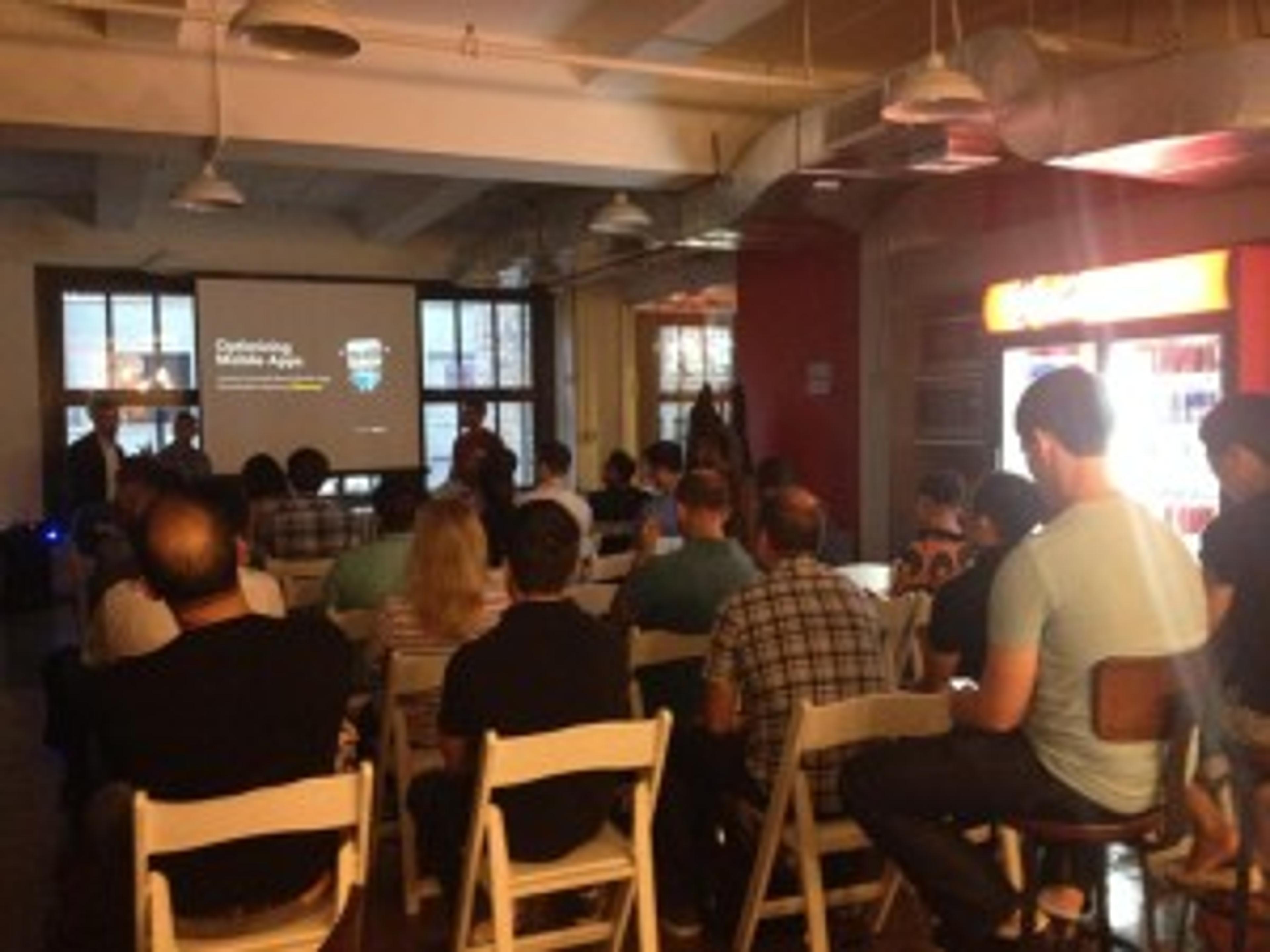 A group of people seated in a room facing a presentation screen. The audience is engaged in a talk about optimizing mobile apps.