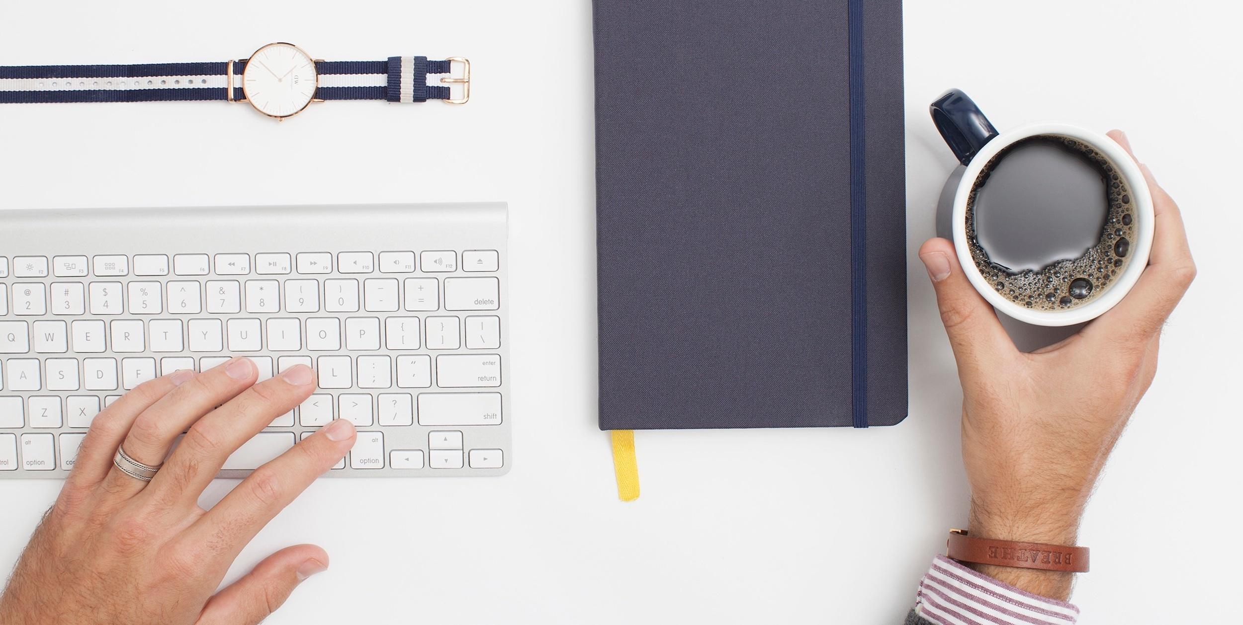 A person's hand typing on a keyboard next to a notebook and a cup of coffee. A watch is also visible on the table.