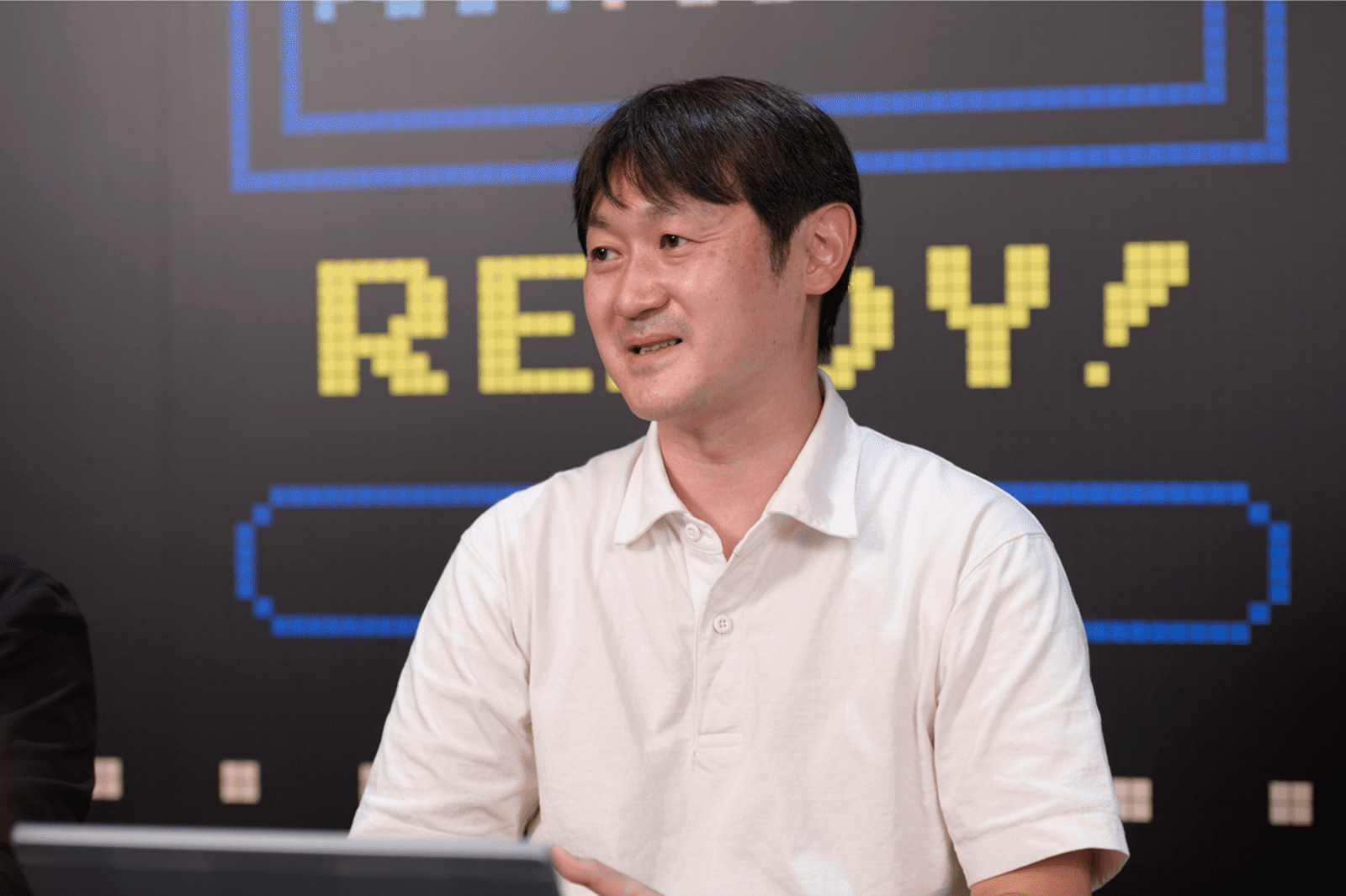バンダイナムコ社員の一人が、ゲーム画面の背景の前で腕をくんで座ってインタビューに答えている様子