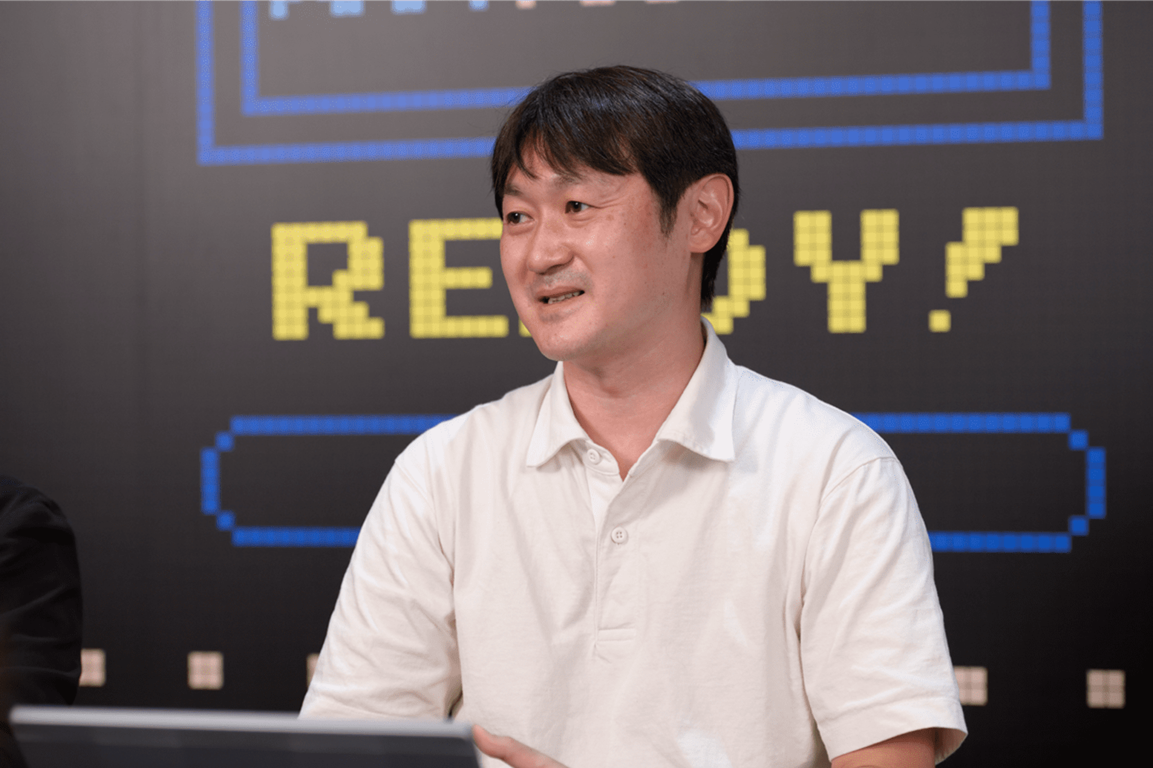 バンダイナムコ社員の一人が、ゲーム画面の背景の前で腕をくんで座ってインタビューに答えている様子