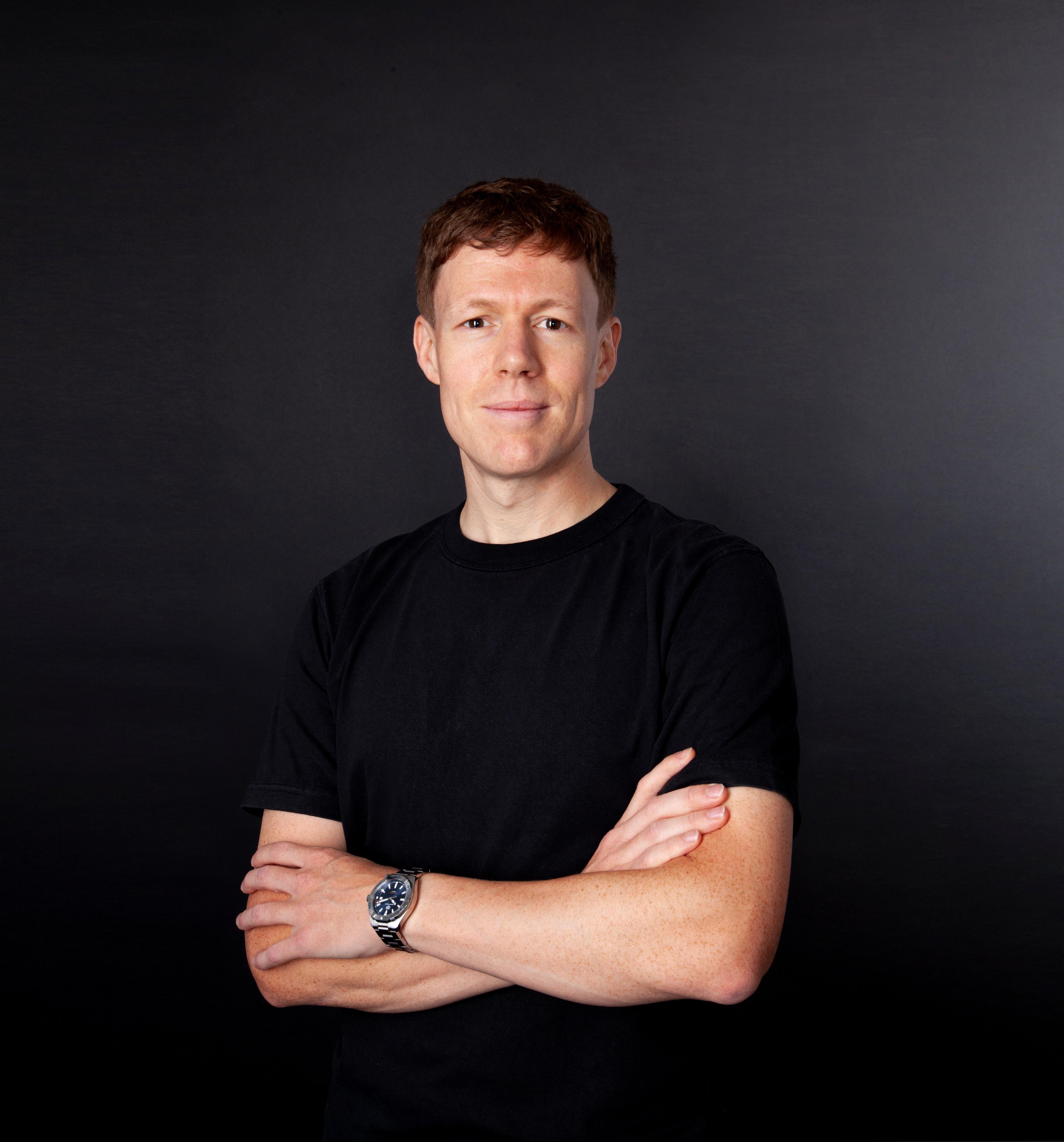 A man with short hair stands confidently with his arms crossed, wearing a black t-shirt against a dark background. He has a watch on his left wrist.
