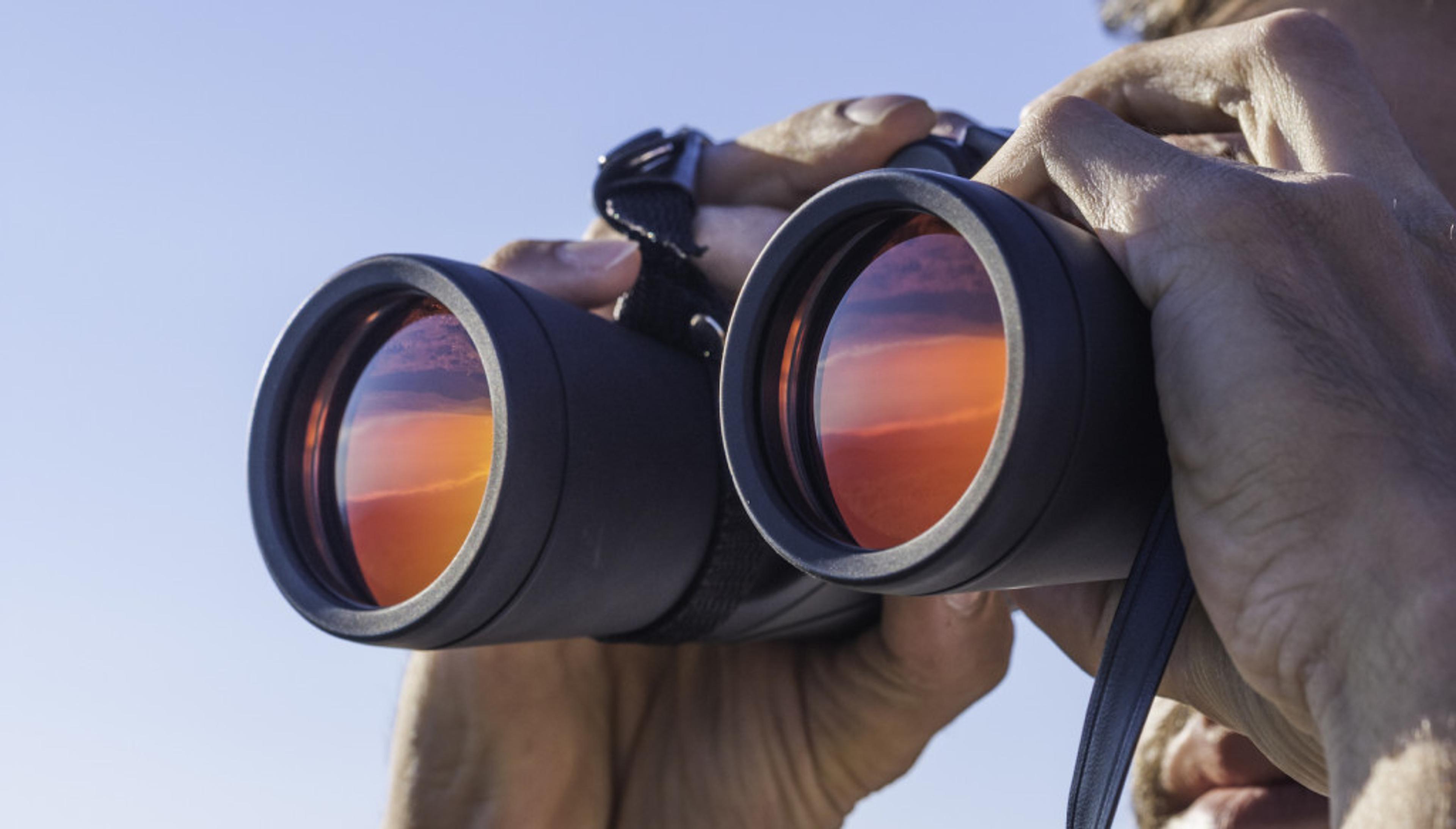 A person holds binoculars up to their eyes, with a sunset reflected in the lenses. The background is a clear blue sky.