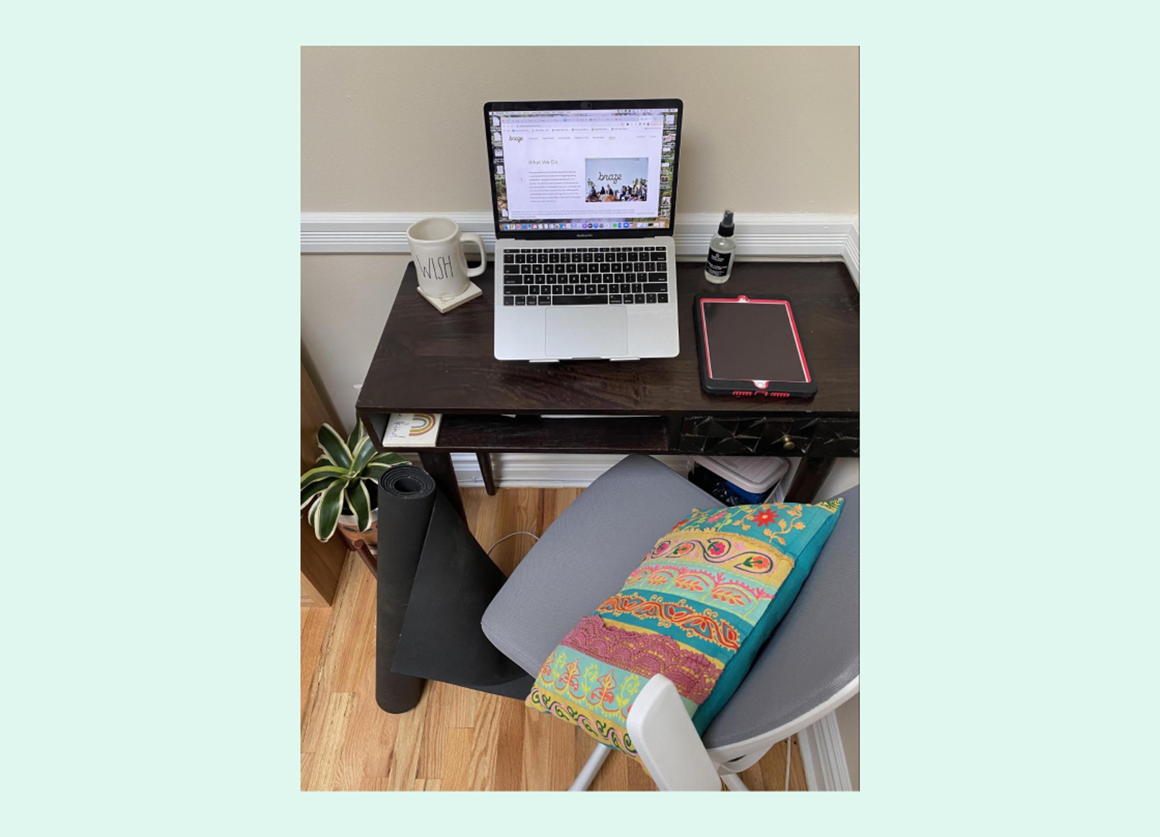 A desk setup featuring a laptop, a mug, a bottle, and a tablet, with a colorful pillow on a chair and a yoga mat on the floor. A small plant is visible in the corner.