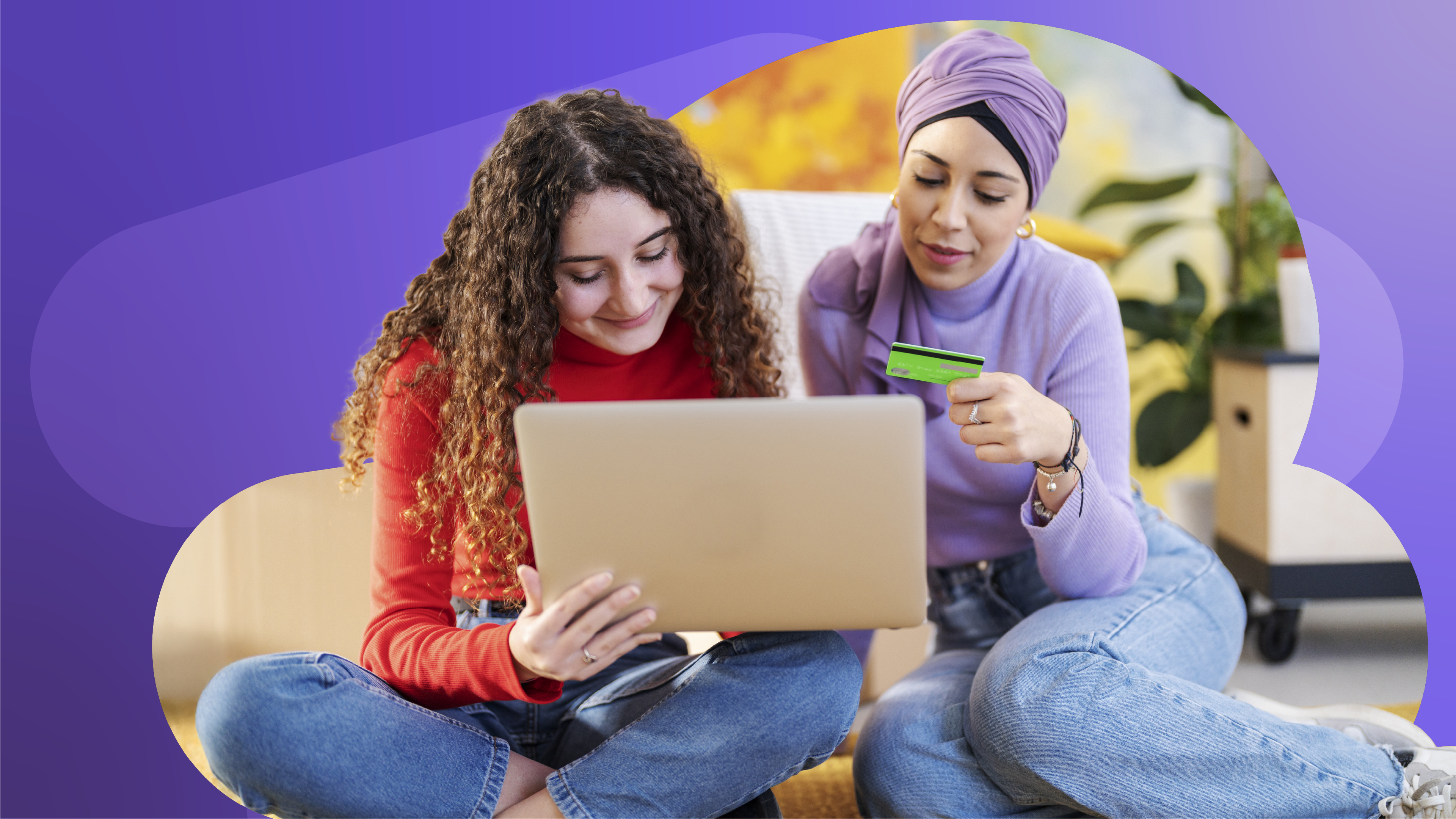 two women are sitting on the floor looking at a laptop and one is holding a credit card