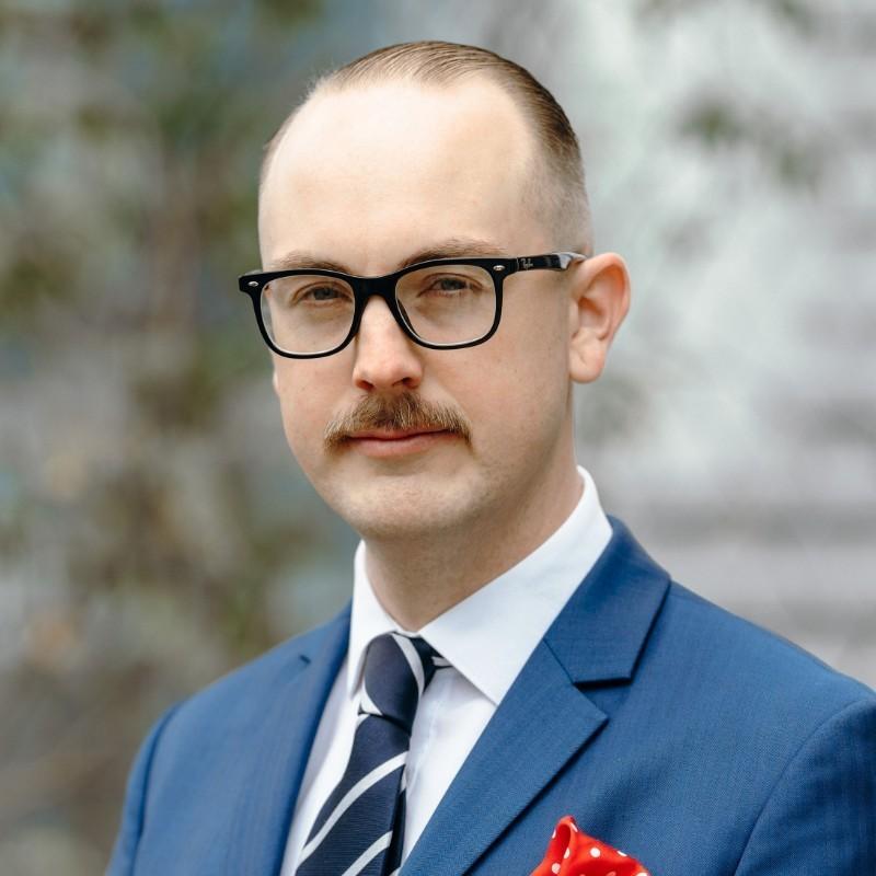 A man in a blue suit and glasses stands outdoors, looking confidently at the camera. He has a mustache and a red pocket square.