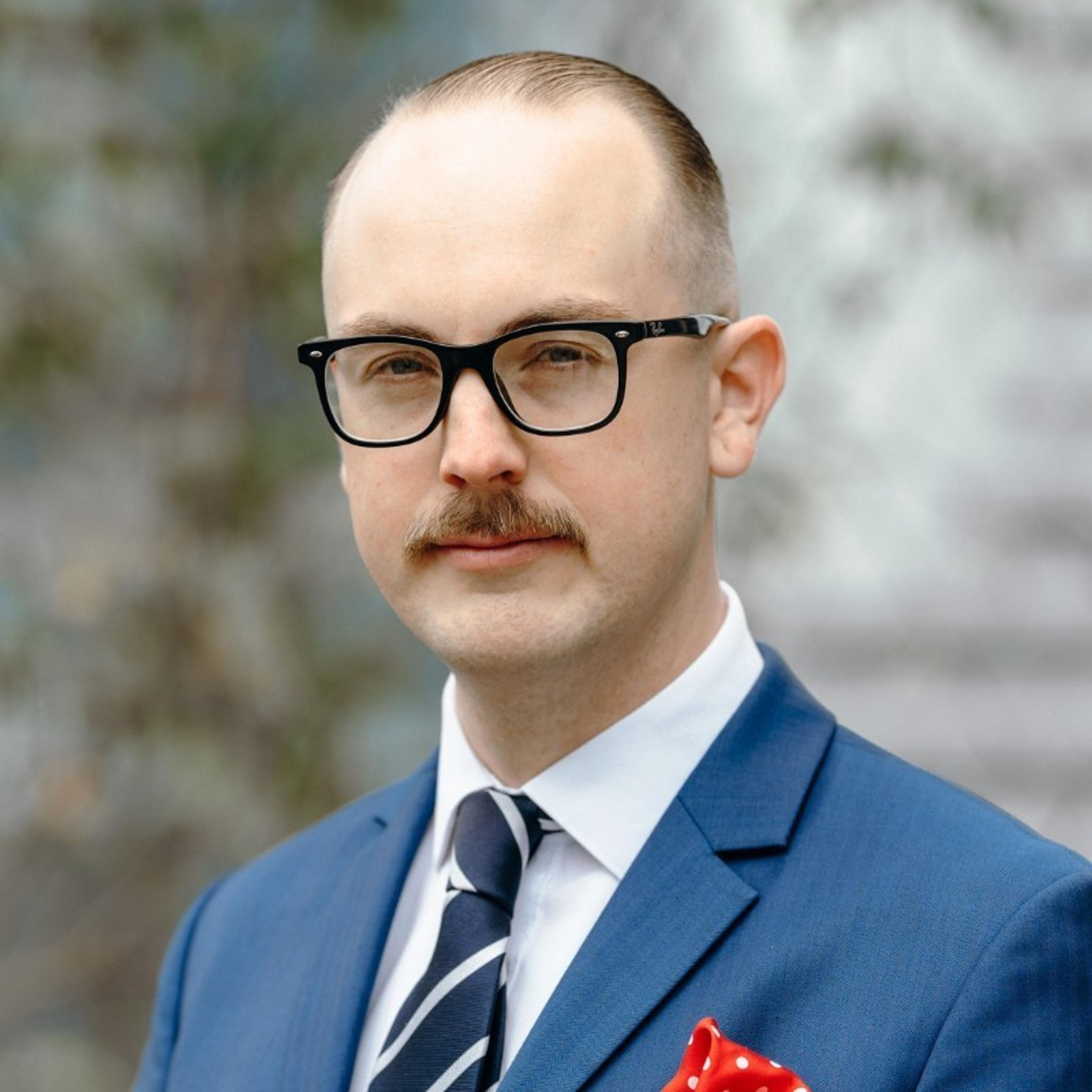 A man in a blue suit and glasses stands outdoors, looking confidently at the camera. He has a mustache and a red pocket square.