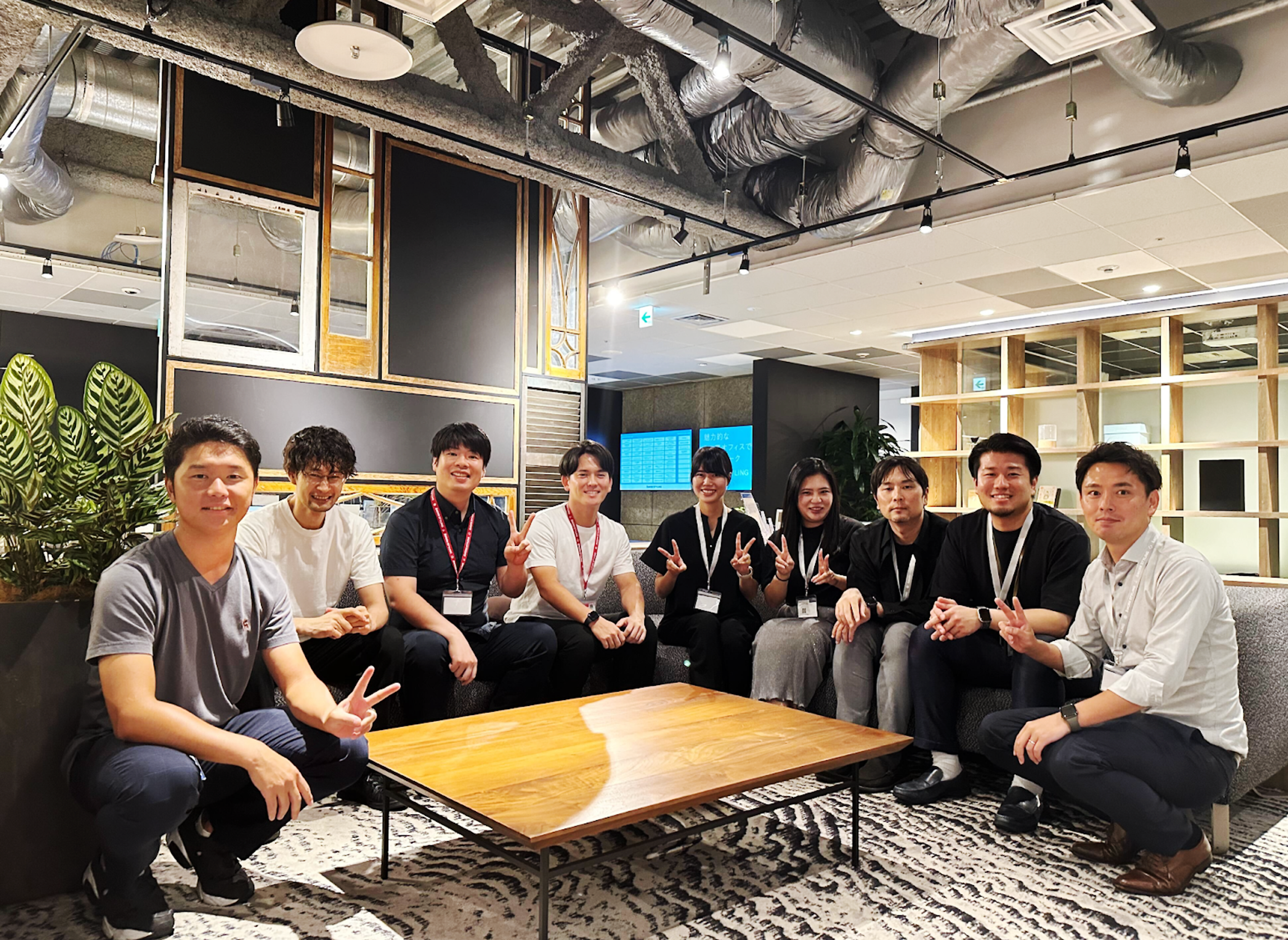 オフィスのラウンジでポーズをとる9人の若者たちが写っています。背景には観葉植物とモダンなインテリアがあります。