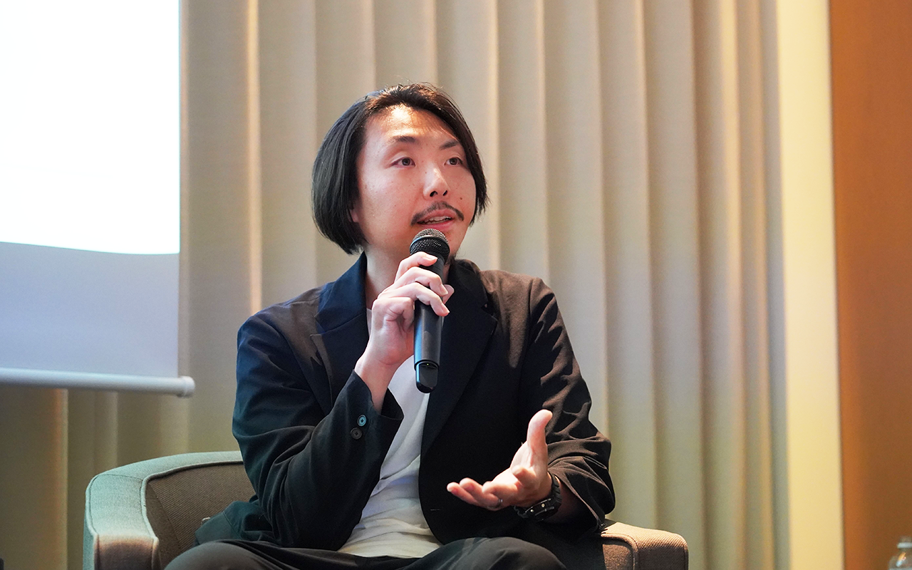 マイクを持って話す男性が座っている様子。背景にはカーテンとスクリーンが見える。