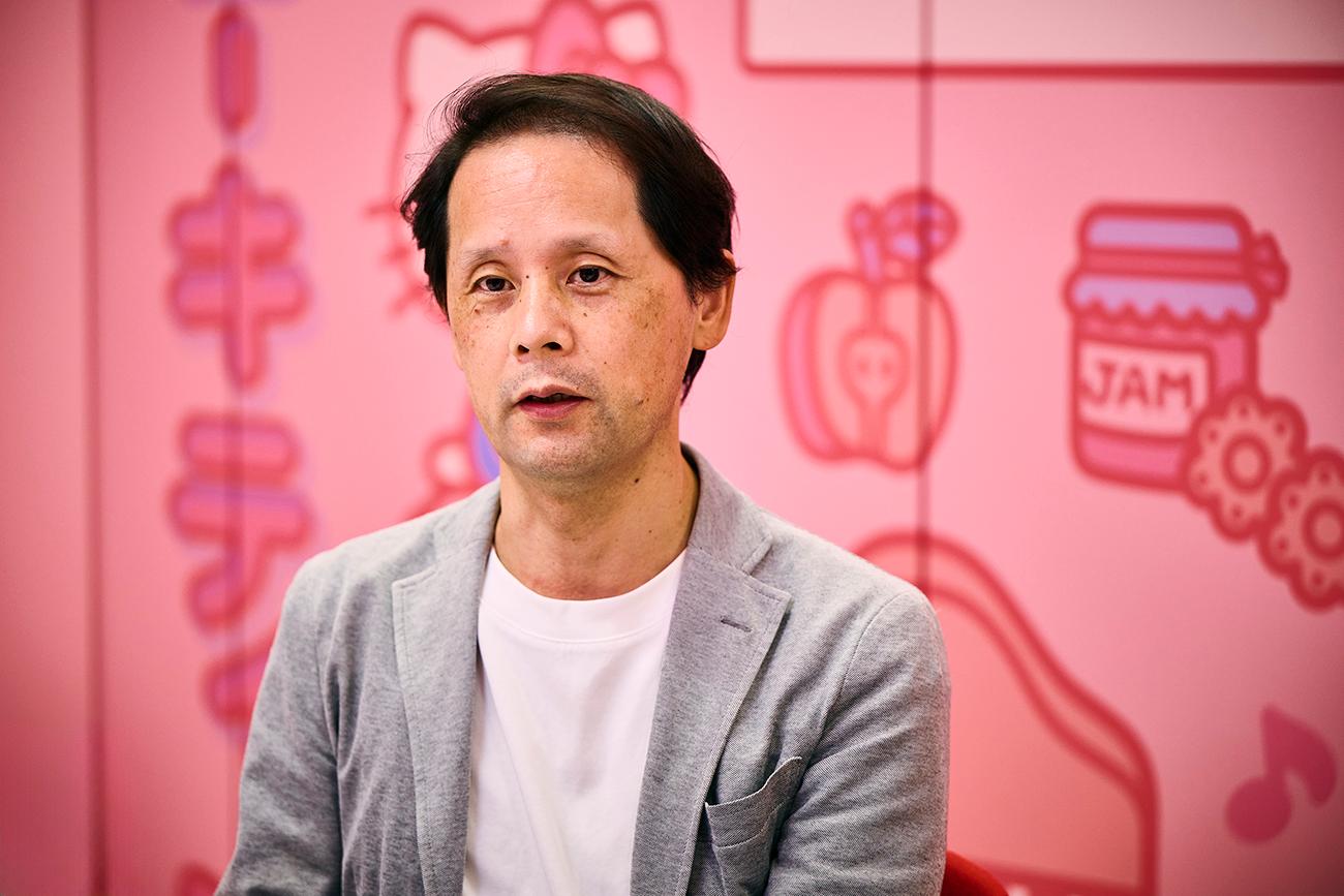 灰色のジャケットを着た男性(サンリオ デジタル事業本部 デジタル事業開発部 CX推進課 髙松 貴宏氏)が、ハローキティやその他の漫画のイラストで飾られたピンクの壁の前に座っている。