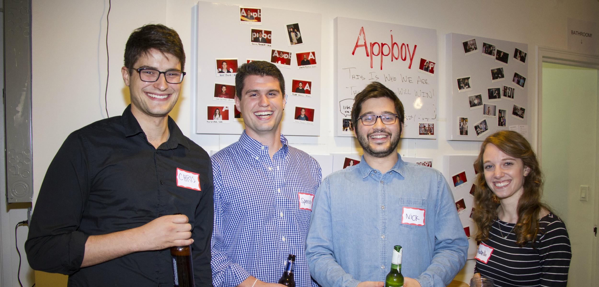 Four people stand together smiling at a social event, with a wall behind them displaying photos and text. They are holding drinks and wearing name tags.