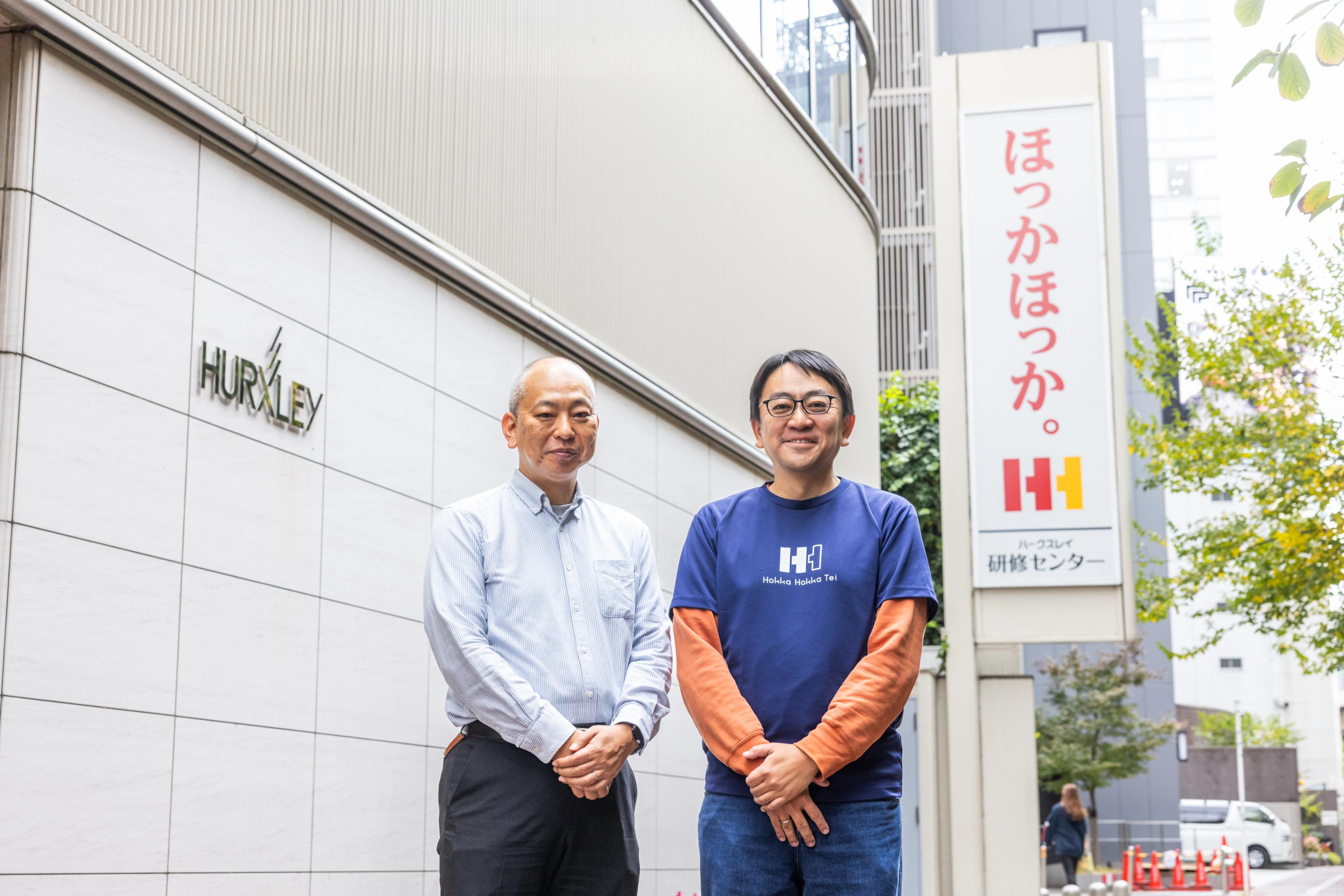 二人の男性が「ほっかほっか」と日本語の看板がある建物の外に立っている。