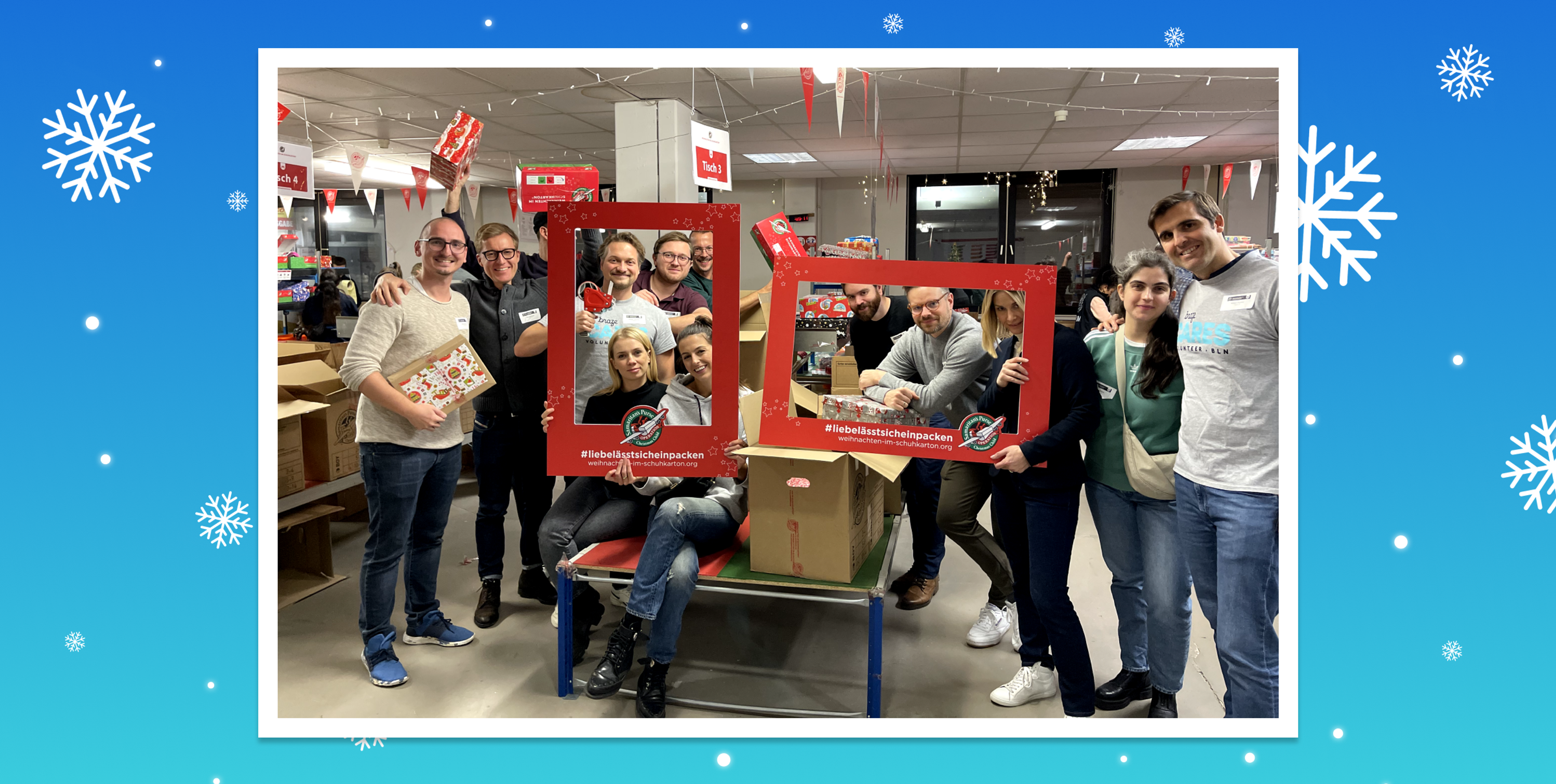 A group of people poses together with festive decorations, holding a large red frame that reads "#TeamChallengeAccepted." They are in a brightly lit room filled with boxes and holiday-themed items.