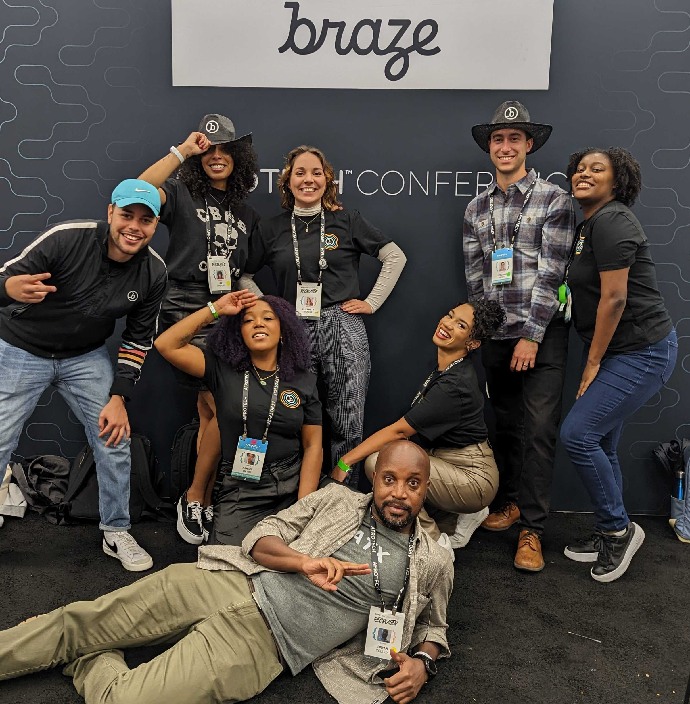 A group of eight people poses together in front of a Braze sign at a conference, with some making playful gestures. The setting is casual and festive.