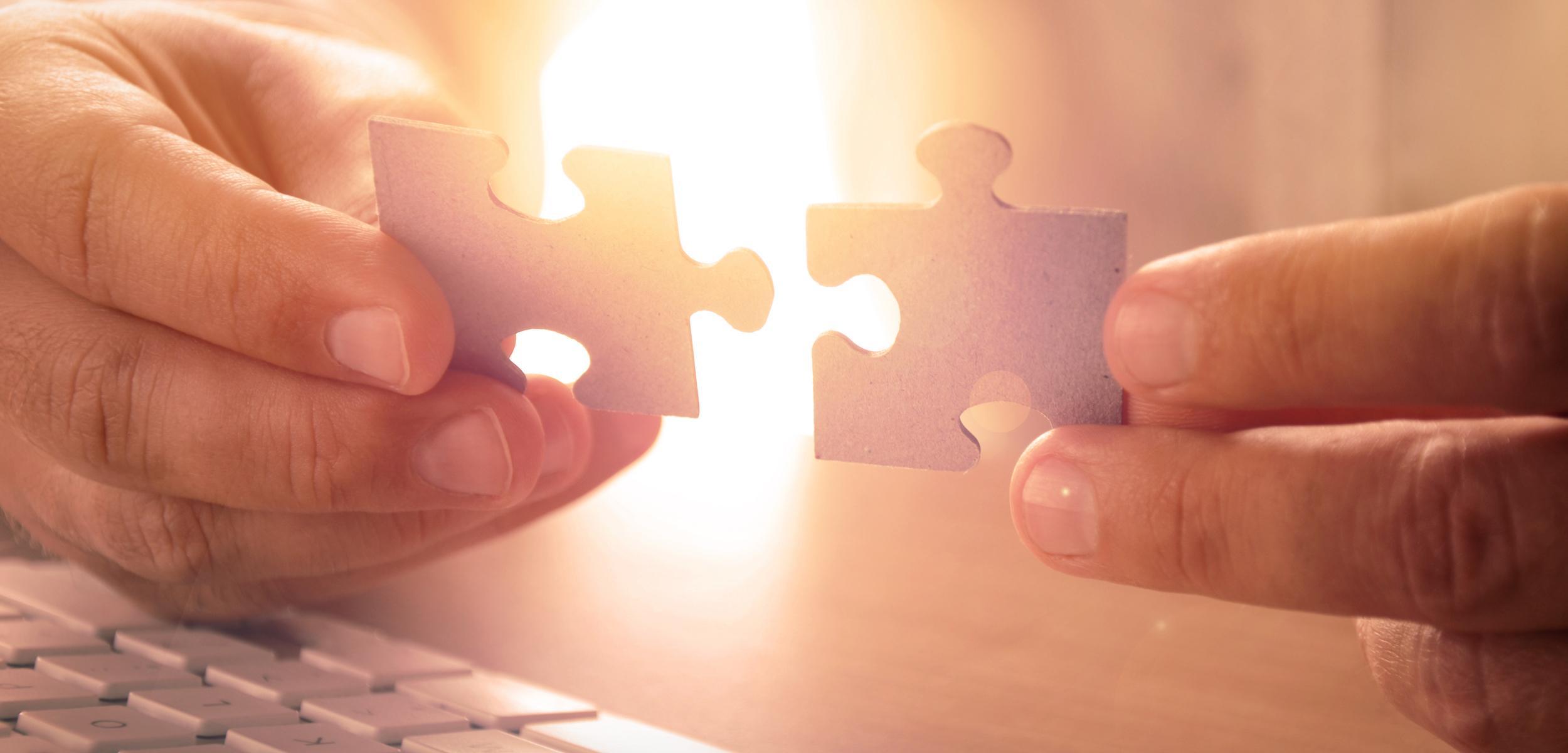 Two hands hold puzzle pieces, about to connect them, with a soft light in the background. A keyboard is partially visible in the foreground.