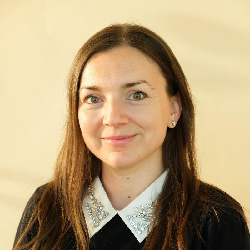 Smiling woman with long brown hair in a black top with a jeweled white collar, posing against a plain light background.