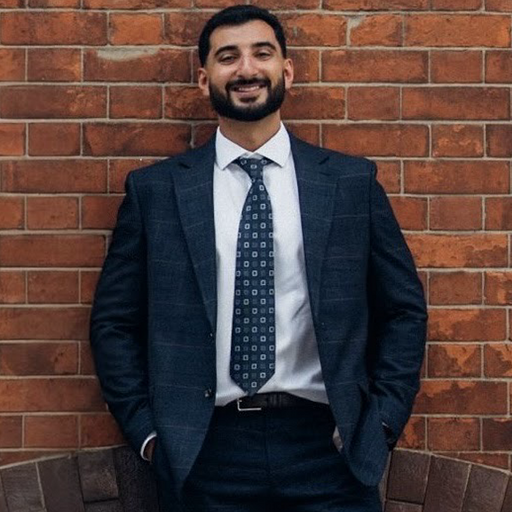 Man in a dark suit and patterned tie standing and smiling in front of a red brick wall.