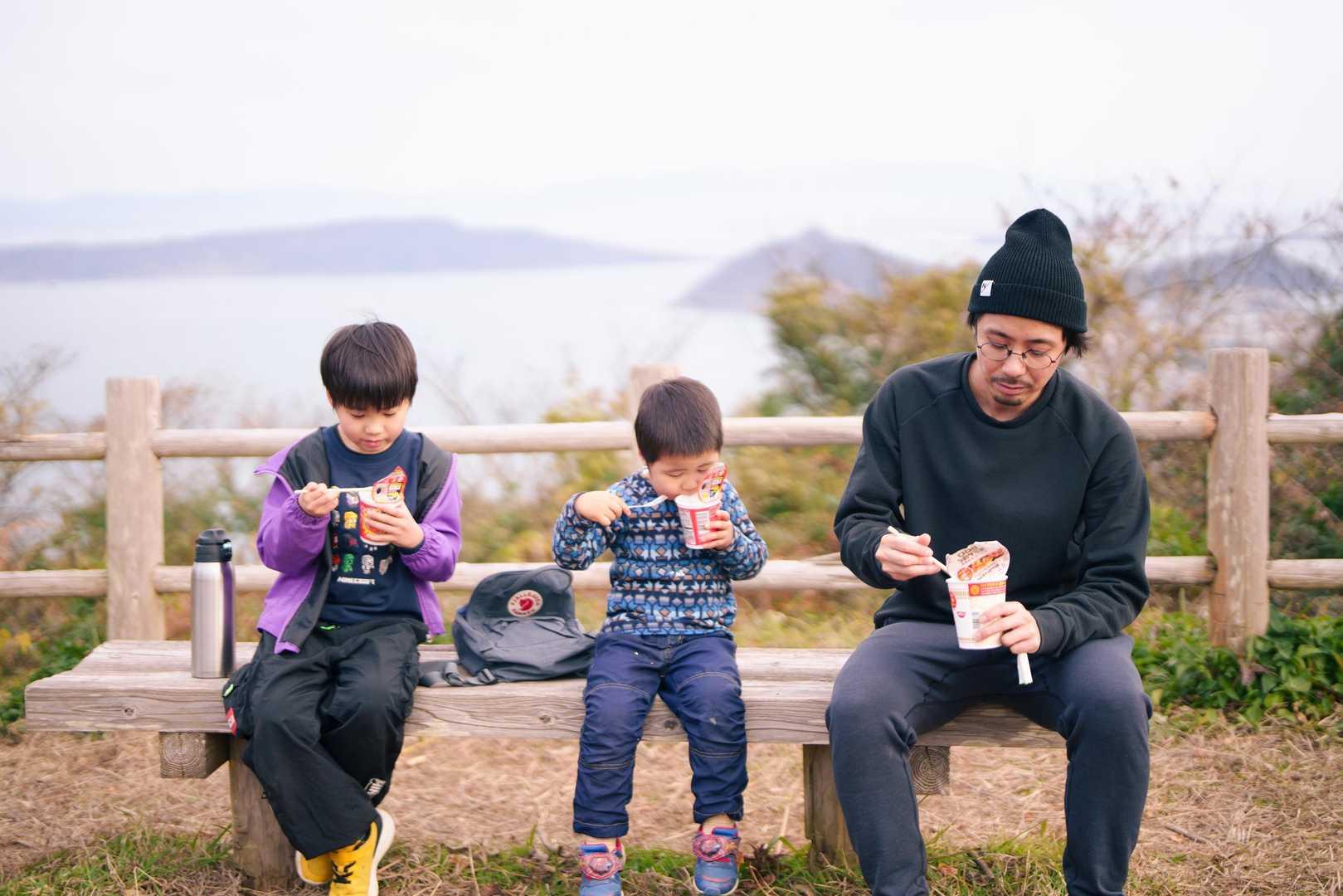 海を望む屋外のベンチに座り、カップ麺を食べる男性と2人の子ども。