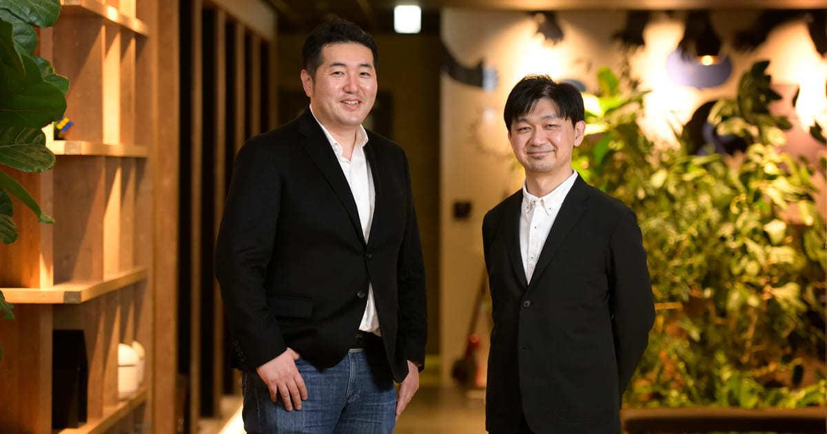 二人の男性がビジネススーツを着て、緑の植物がある室内で立っています。笑顔でカメラを見つめています。