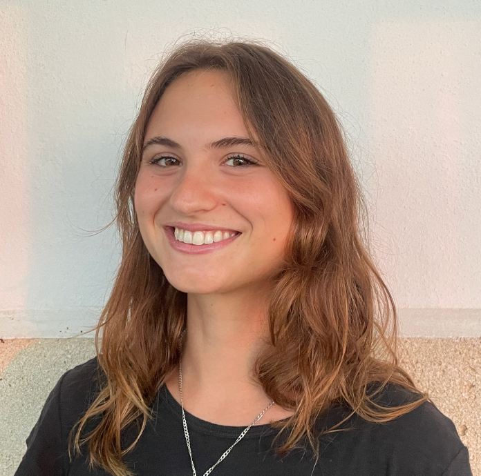A young woman with long, wavy brown hair smiles at the camera, wearing a black shirt. She stands against a light-colored wall.