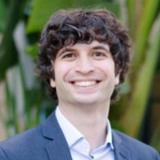 Headshot of a smiling man with curly dark hair wearing a blue suit jacket and light-colored shirt, with greenery in the background.