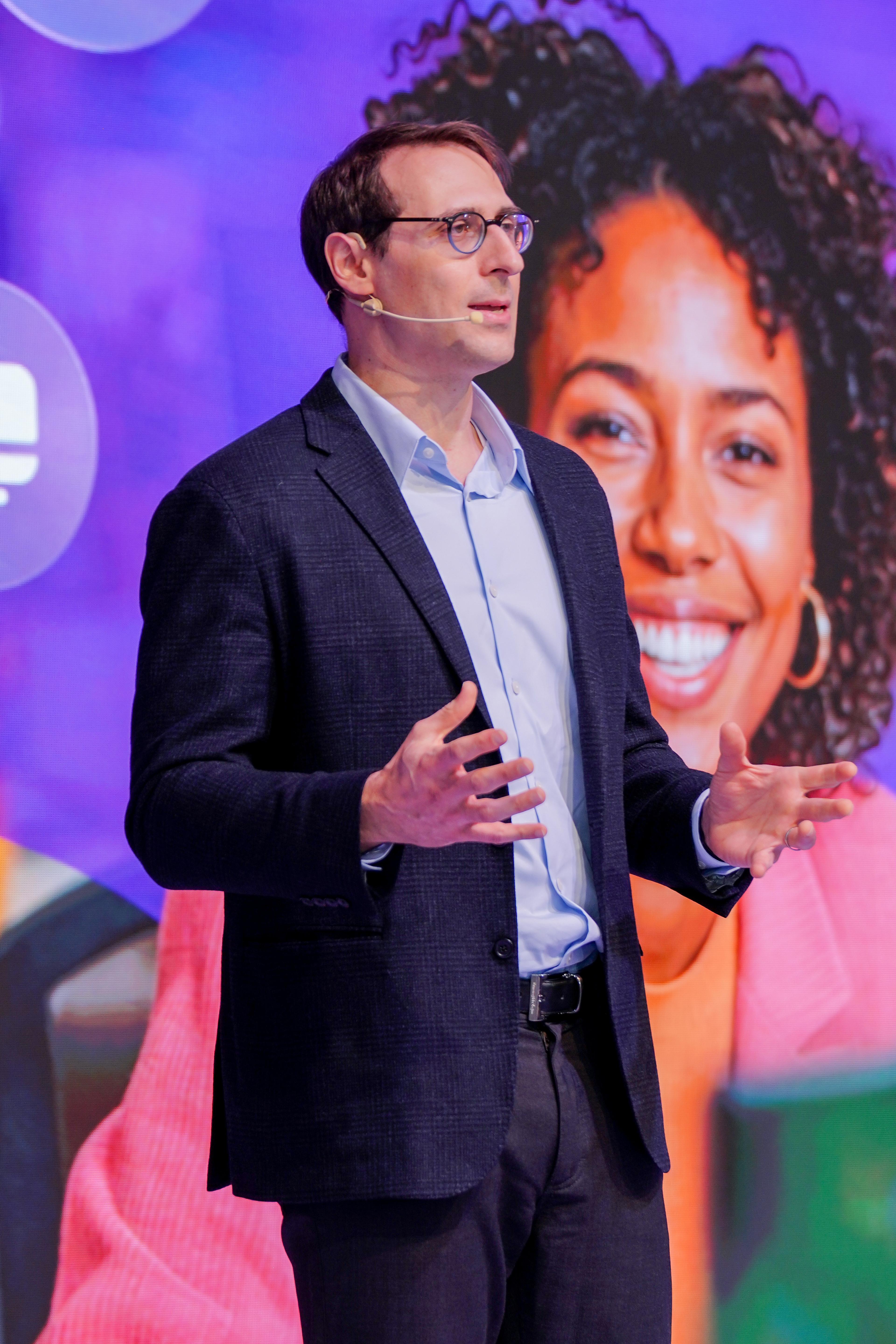 A man in a dark suit jacket and glasses, wearing a headset microphone, speaks against a vibrant background featuring a large image of a smiling woman.