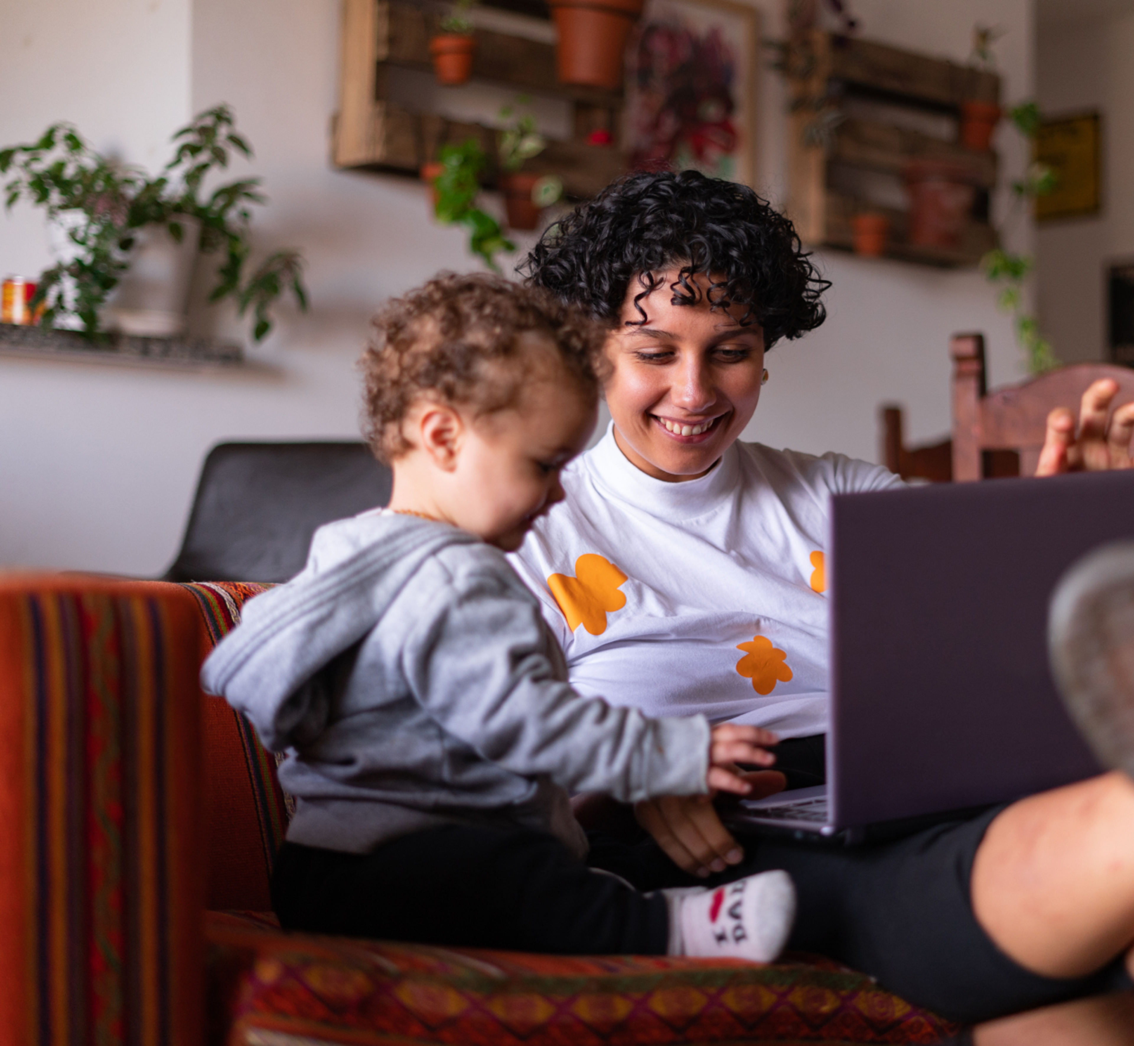 Smiling adult sitting on a couch with a toddler on their lap, both looking at and touching an open laptop in a cozy living room with plants in the background.