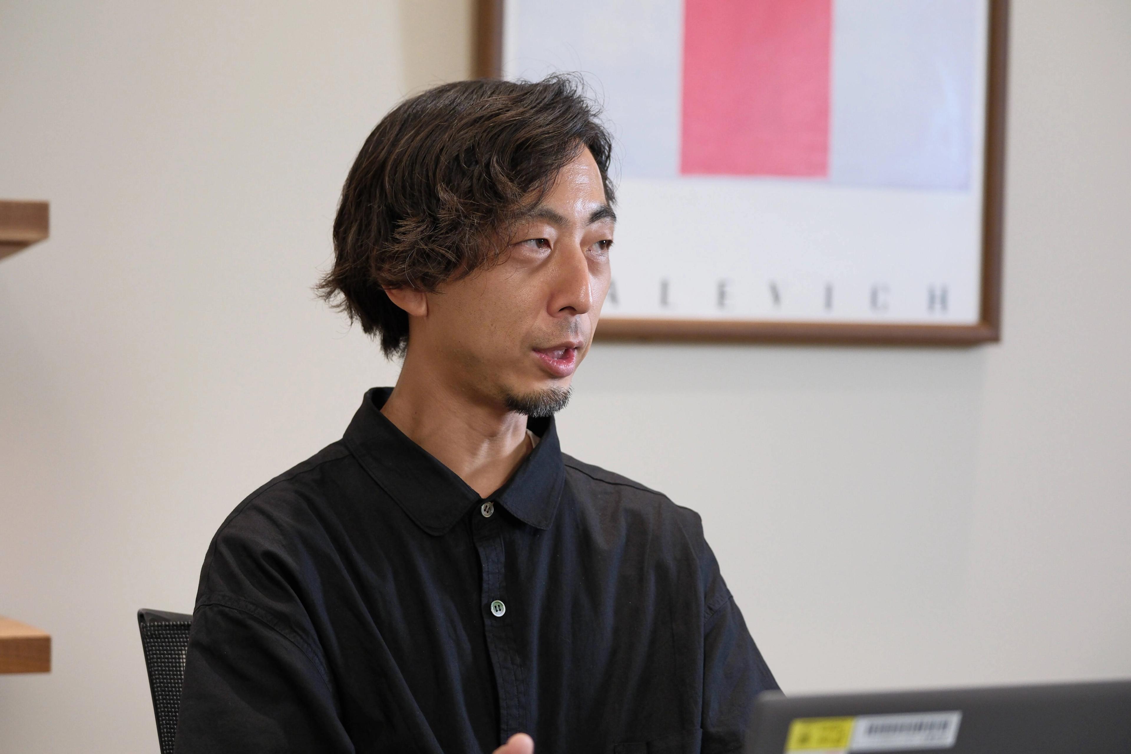 男性が黒いシャツを着て、パソコンの前で話している様子。背景にはアート作品が飾られている。
