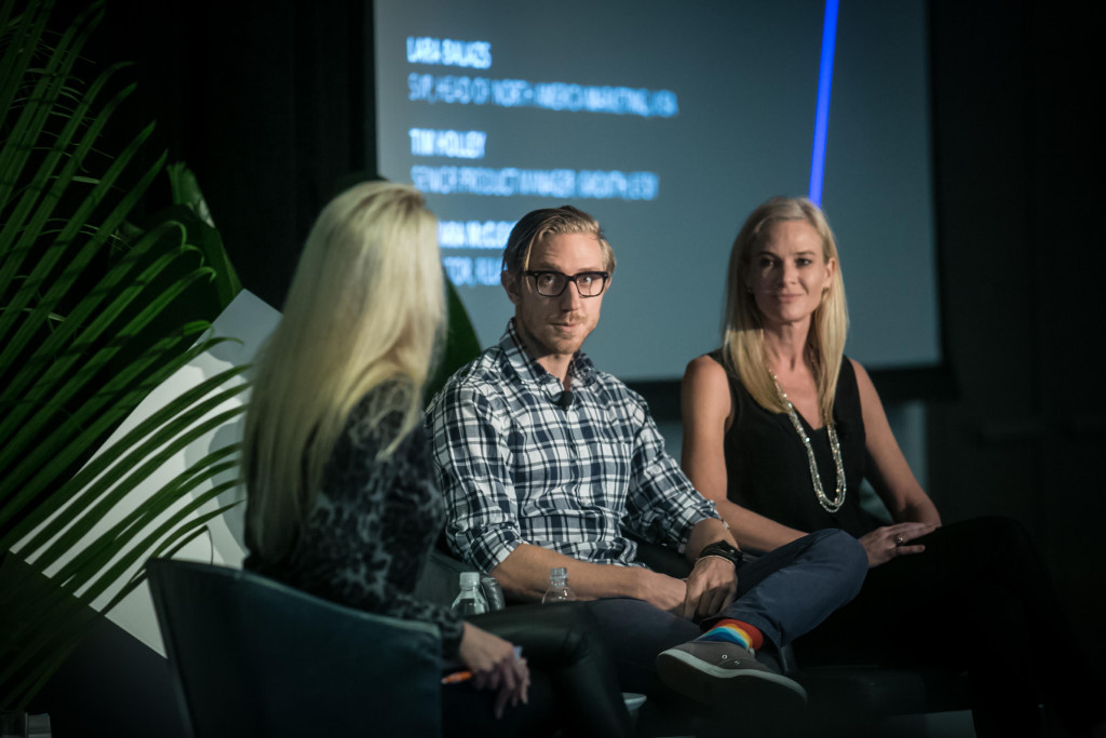 A panel discussion featuring three speakers seated on stage, with one speaker asking questions. The background displays text related to the event.