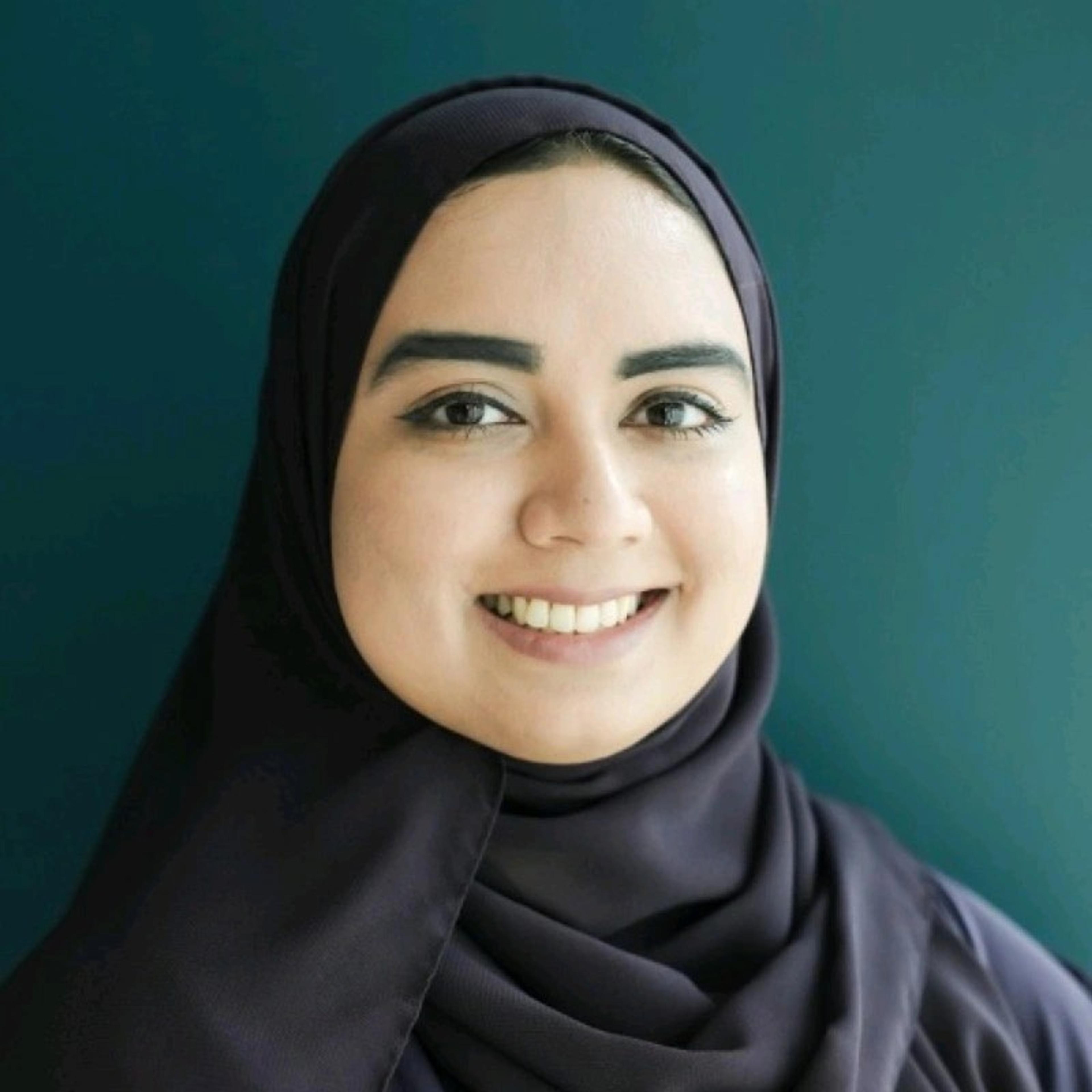 A smiling woman wearing a dark hijab against a teal background. She has bold eyebrows and a friendly expression.