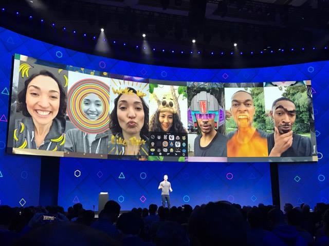 A person stands on stage in front of a large screen displaying various fun and colorful augmented reality filters applied to selfies. The audience is visible in the foreground.