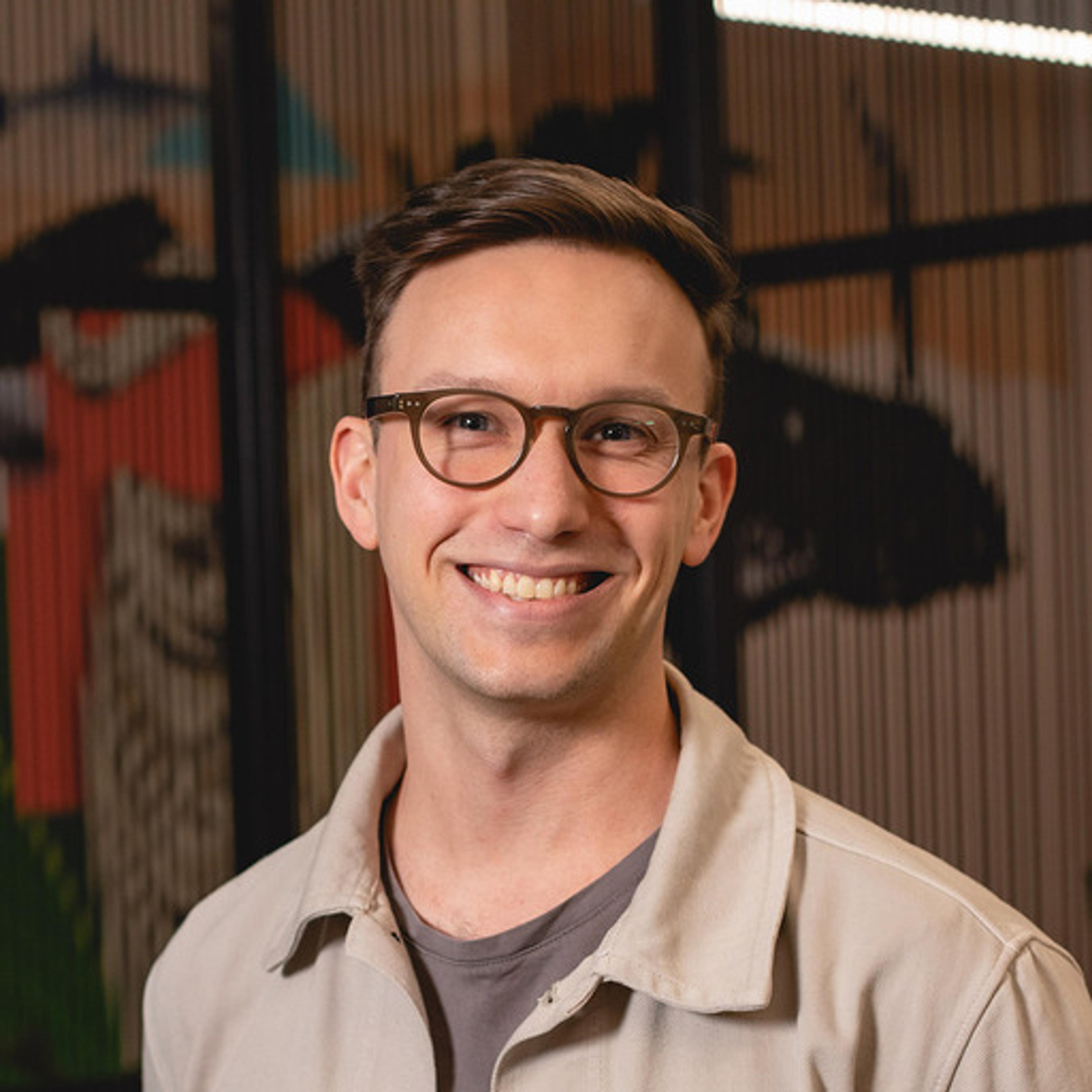 A smiling young man with glasses stands in front of a colorful mural. He is wearing a light-colored jacket over a gray shirt.