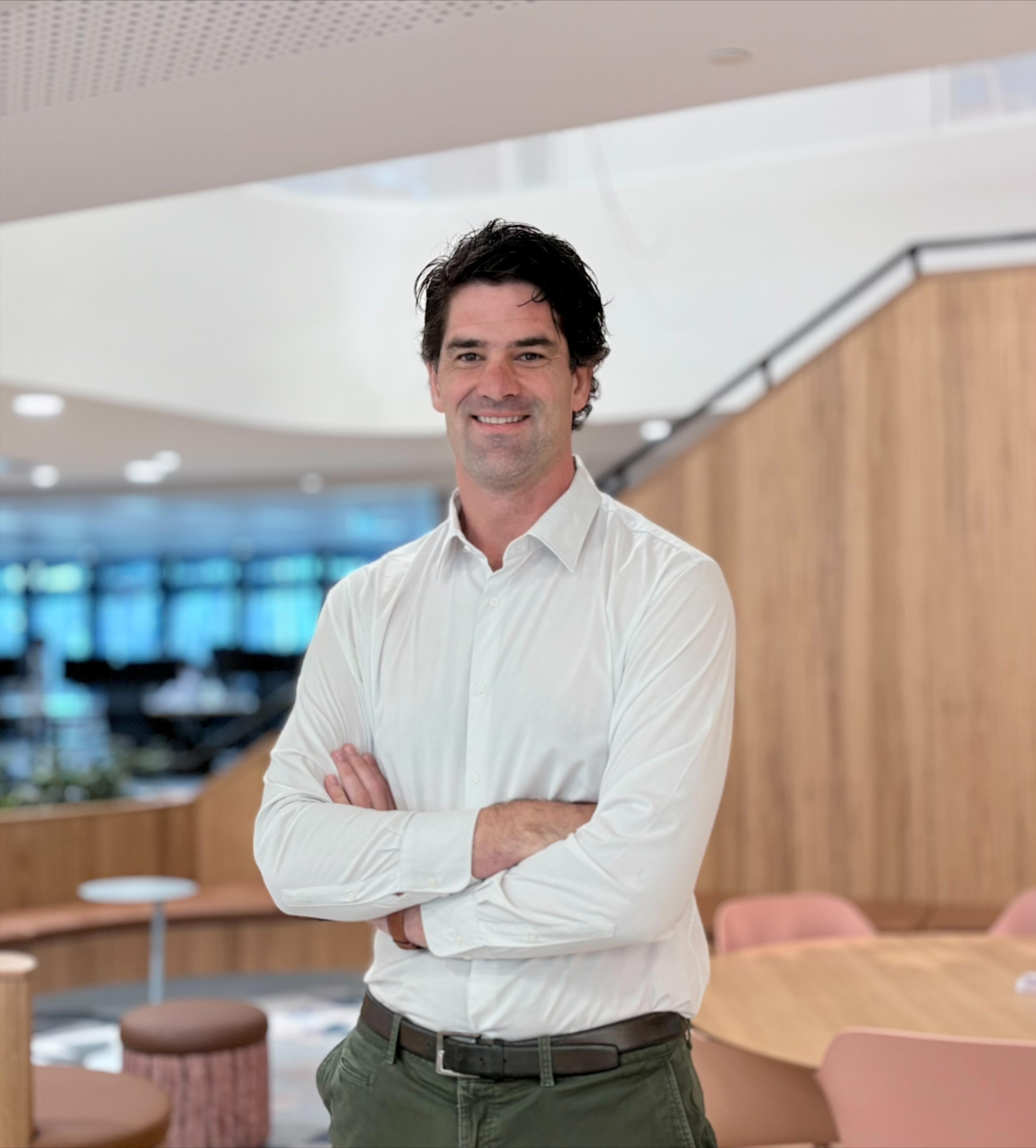 A man in a white shirt stands with his arms crossed, smiling in a modern indoor space with wooden accents and soft seating.
