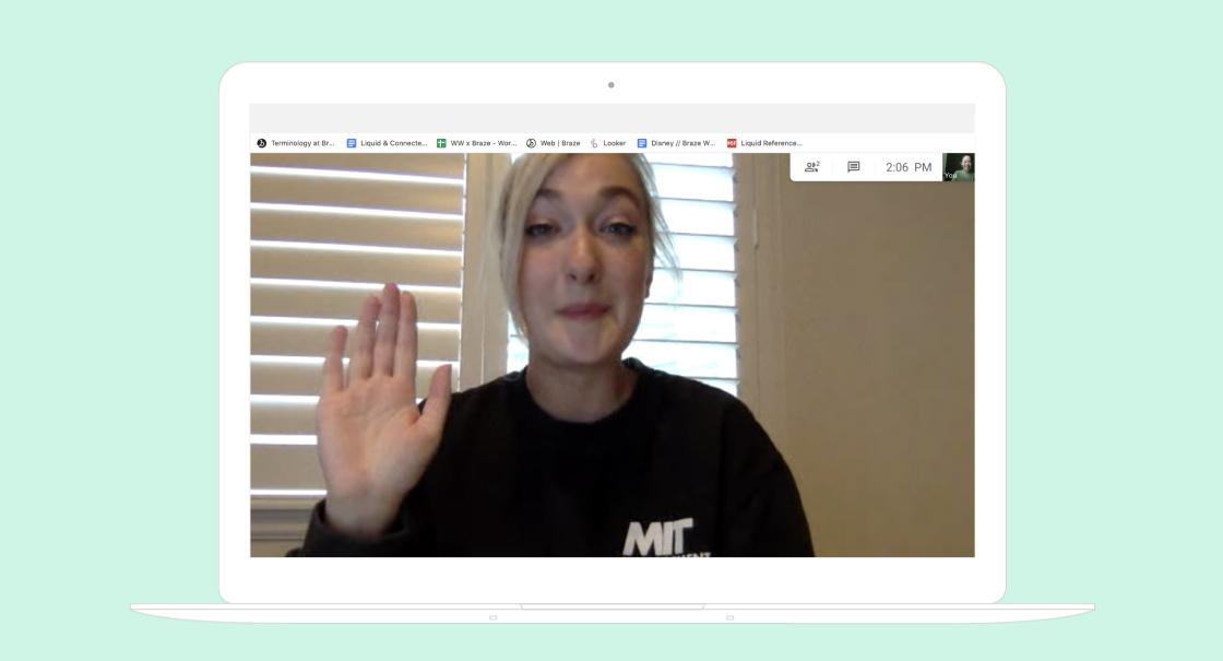 A woman with blonde hair is waving while speaking on a video call. She is wearing a black sweatshirt and appears to be smiling.