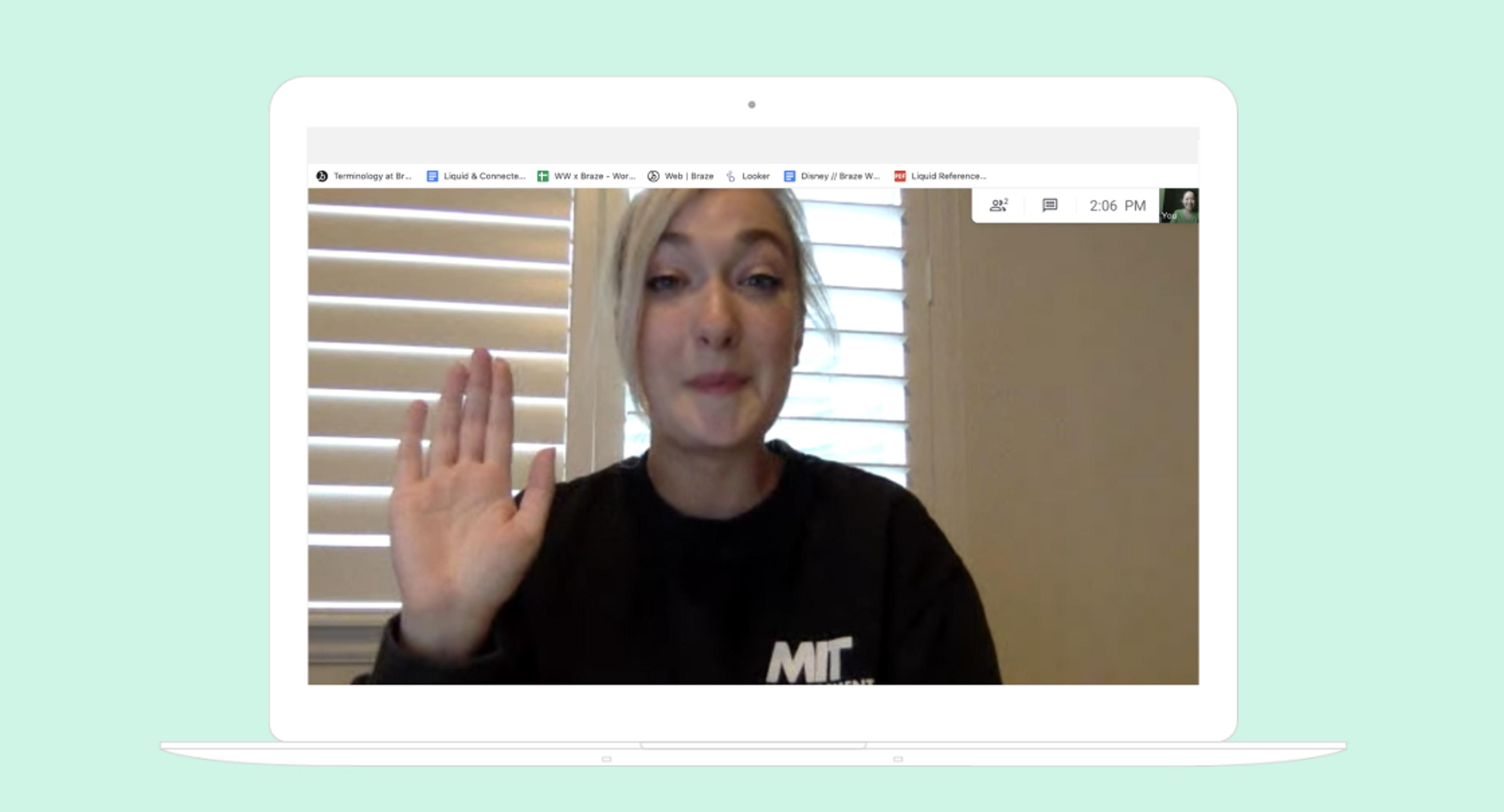 A woman with blonde hair is waving while speaking on a video call. She is wearing a black sweatshirt and appears to be smiling.