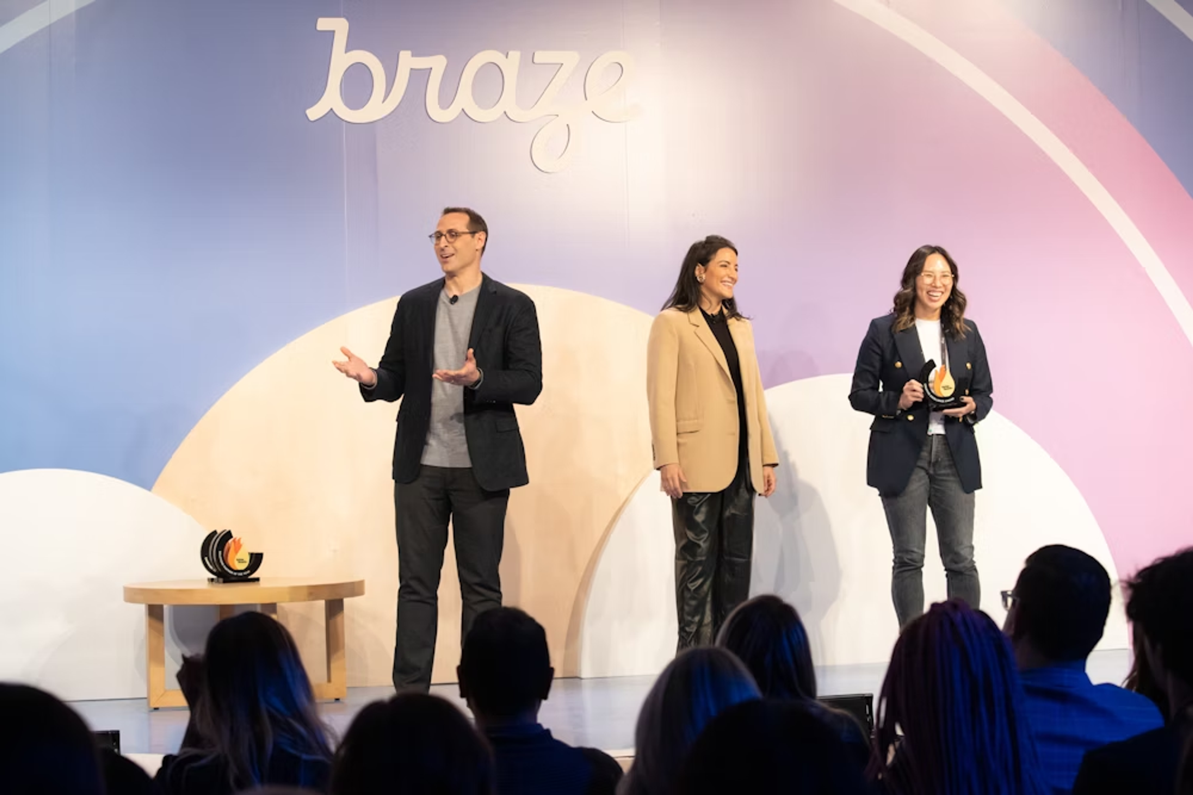 Tres presentadores en un escenario durante un evento, con el logo de "braze" en el fondo. La audiencia está sentada frente a ellos.