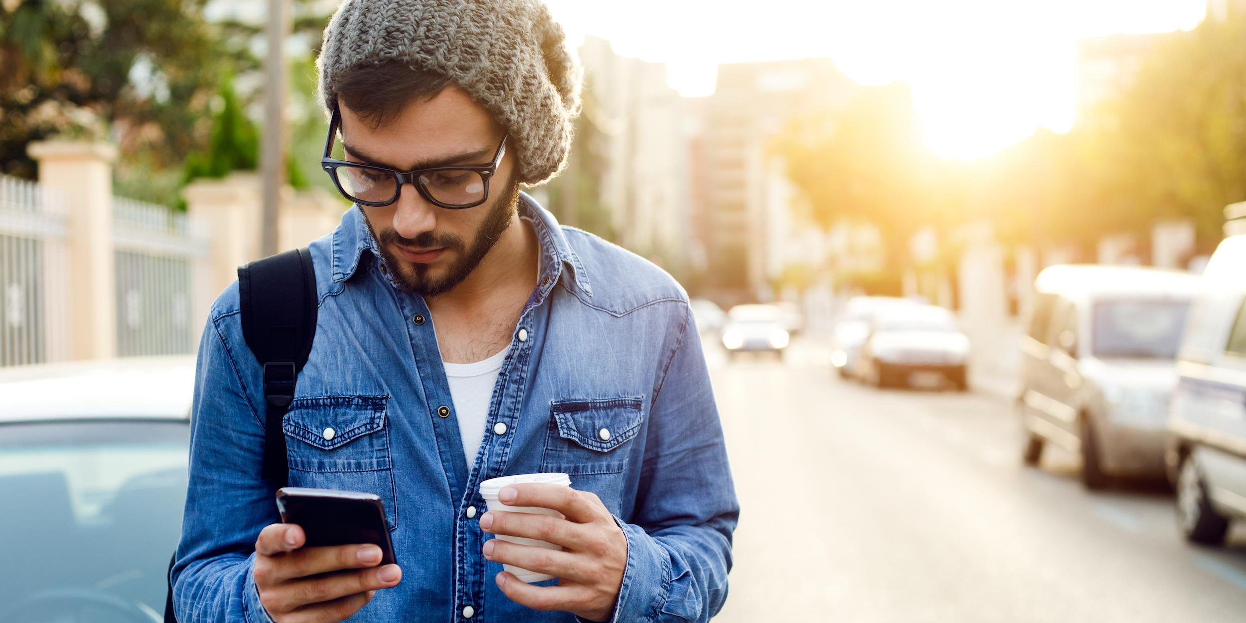 A young man wearing a beanie and glasses stands on a street, looking at his phone while holding a coffee cup. The sun is setting in the background.