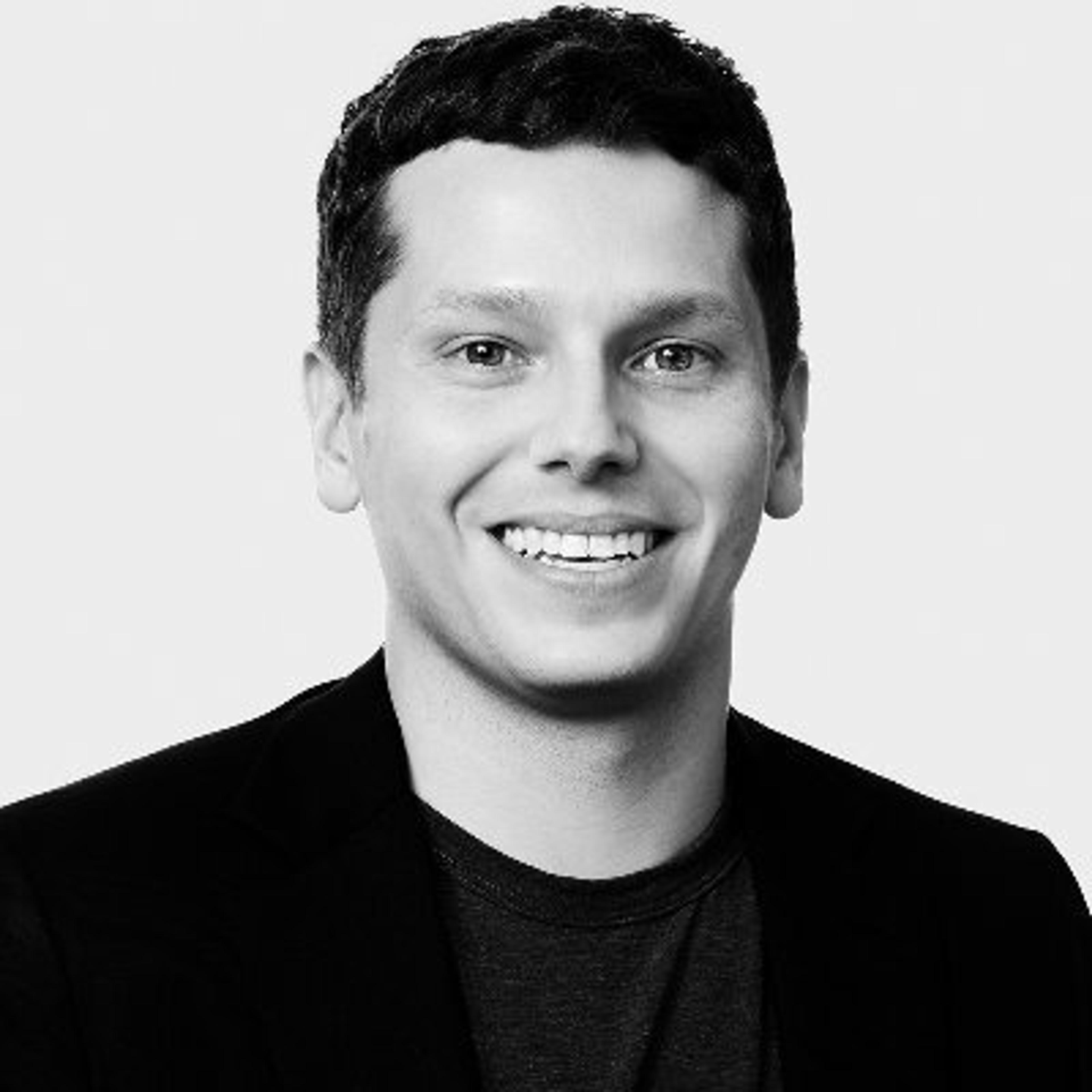 Retrato em preto e branco de um homem sorridente, com cabelo curto e vestindo um blazer. O fundo é claro e simples.