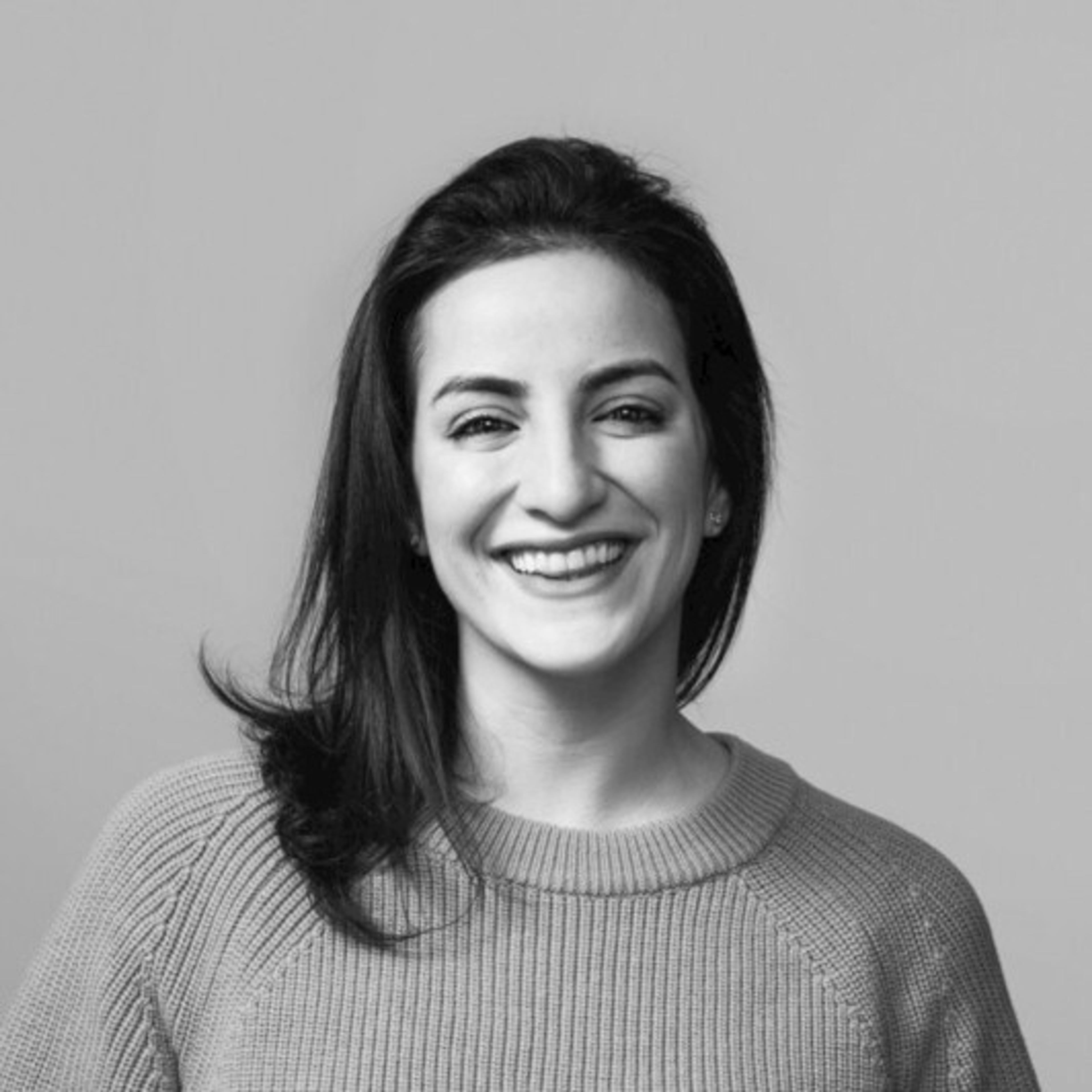 Mulher sorridente com cabelo solto, usando um suéter. O fundo é neutro e claro.