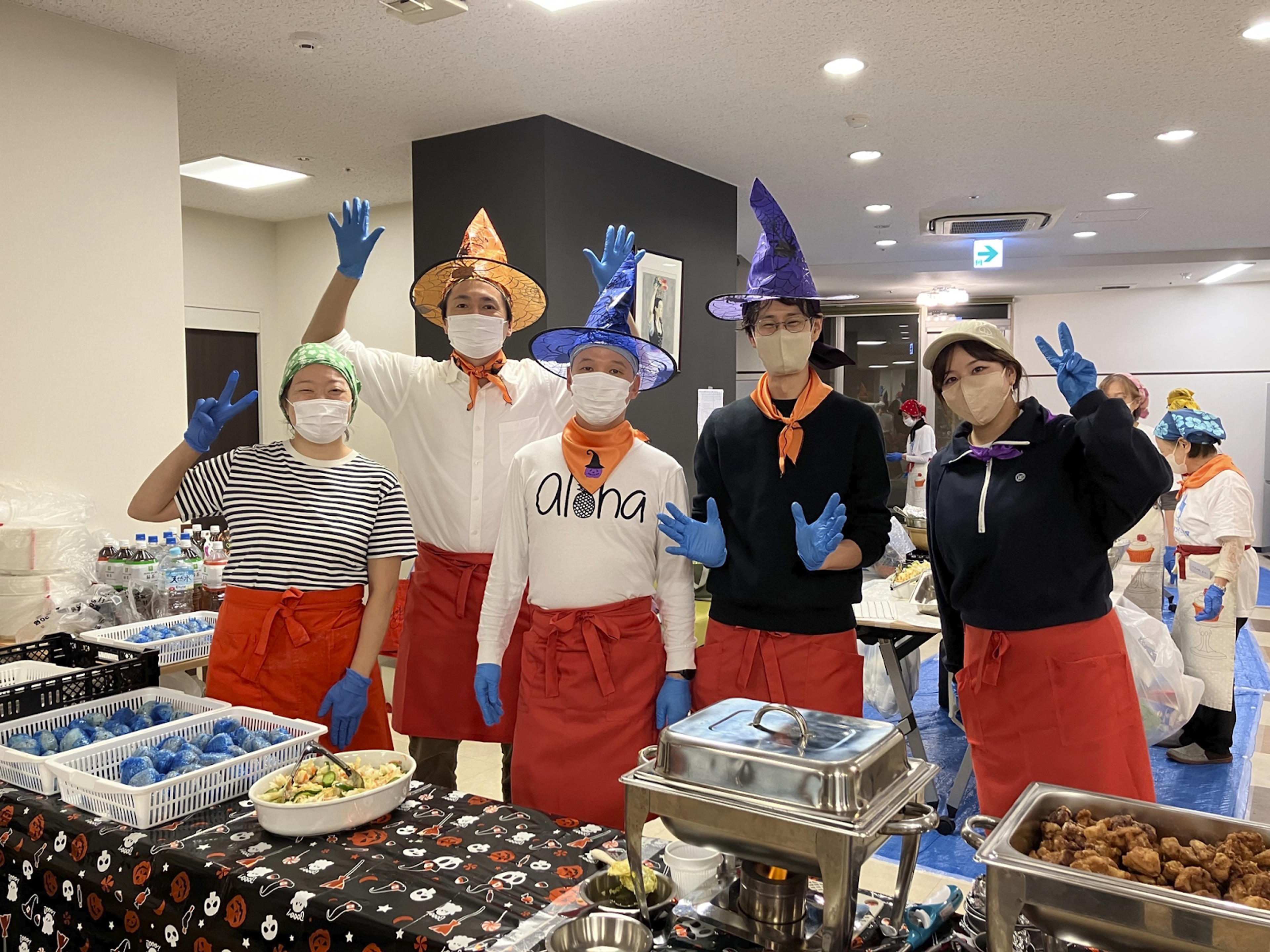 Six people in witches hats, masks, and blue gloves stand behind a Halloween-themed food table, gesturing hello.