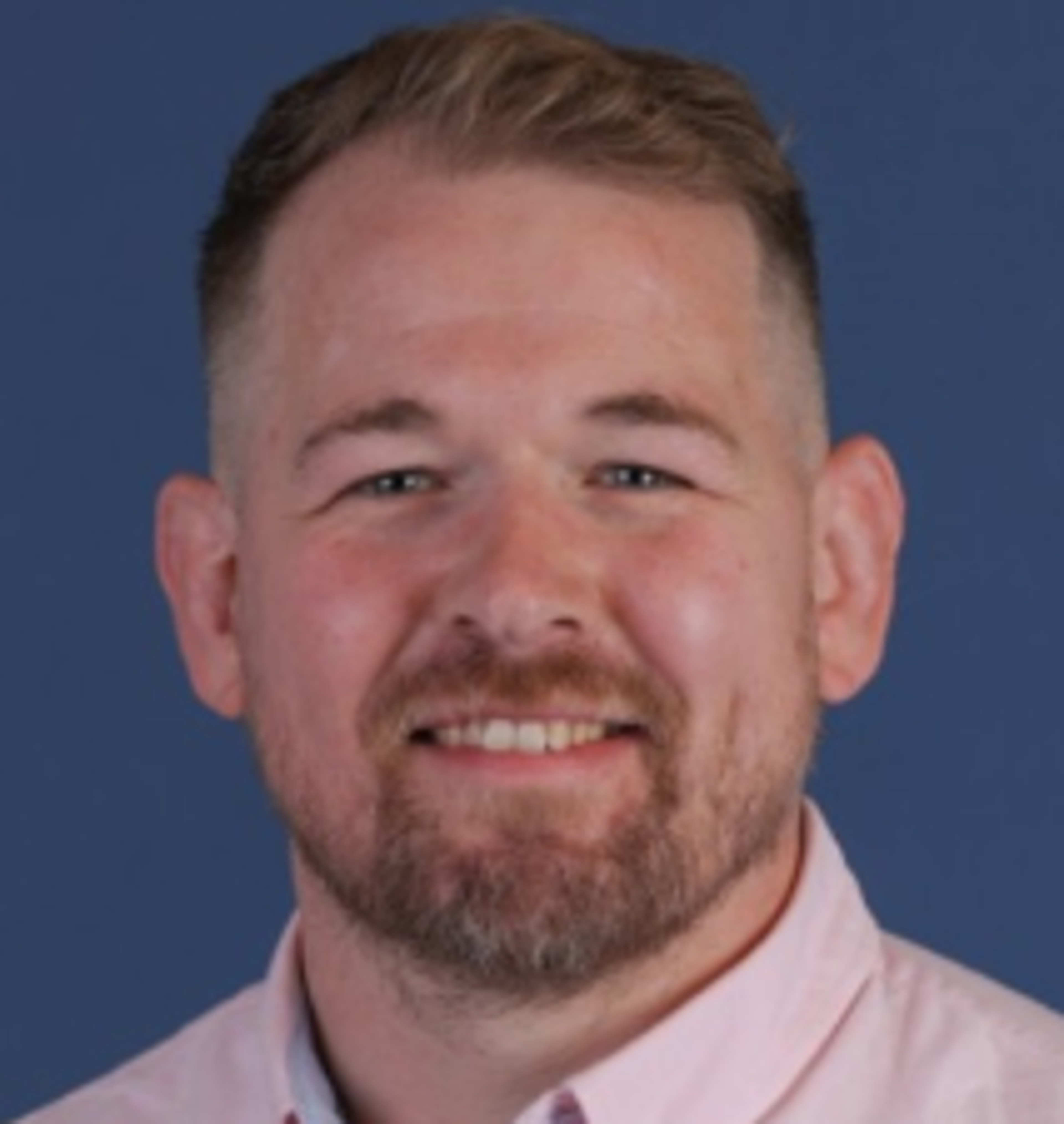 A smiling man with short hair and a beard, wearing a light pink shirt, against a blue background.