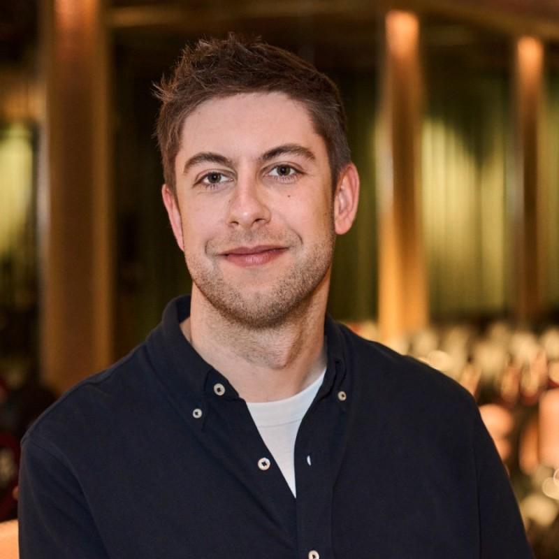 A man with short, light brown hair smiles at the camera, wearing a dark shirt over a light t-shirt. The background features blurred indoor lighting and seating.