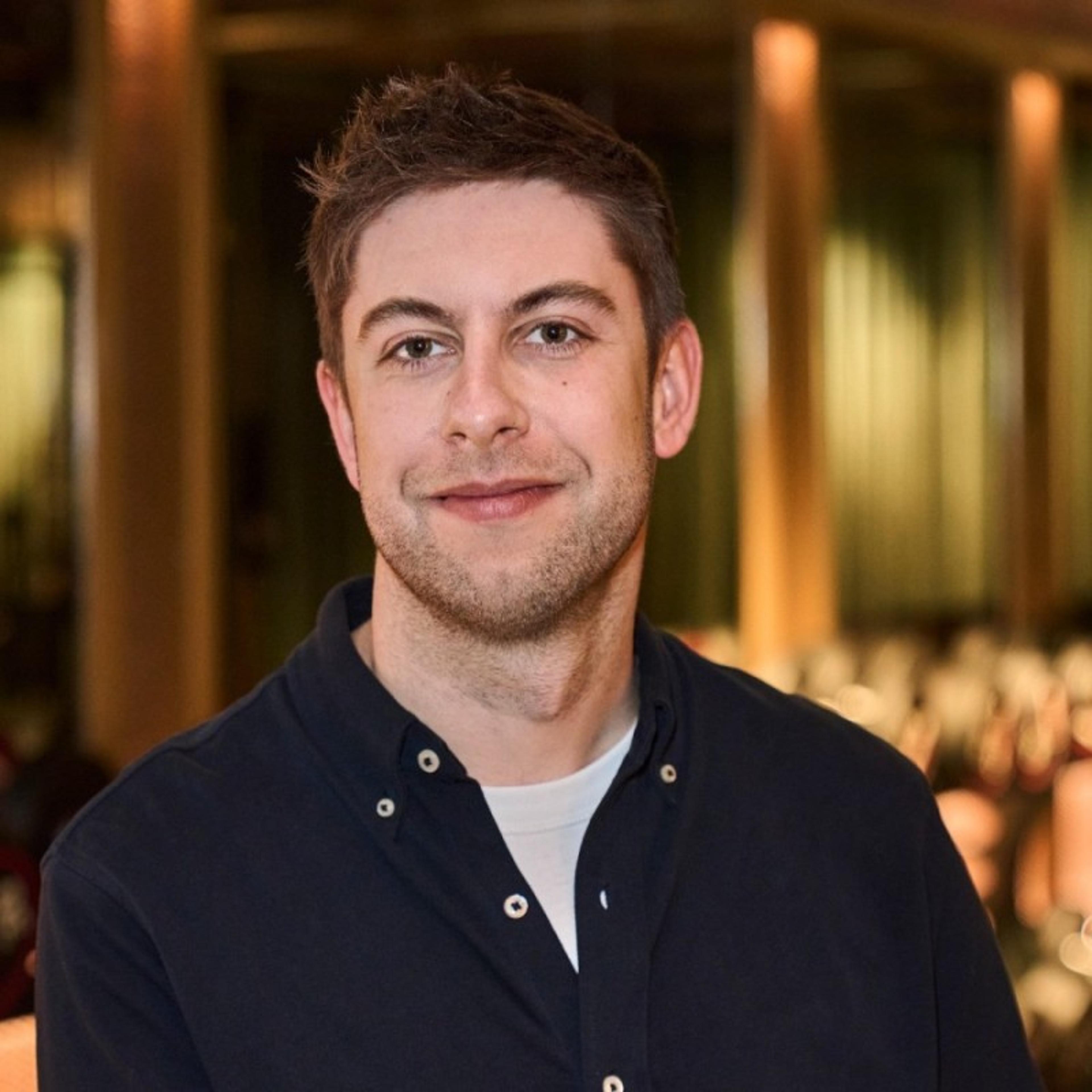 A man with short, light brown hair smiles at the camera, wearing a dark shirt over a light t-shirt. The background features blurred indoor lighting and seating.