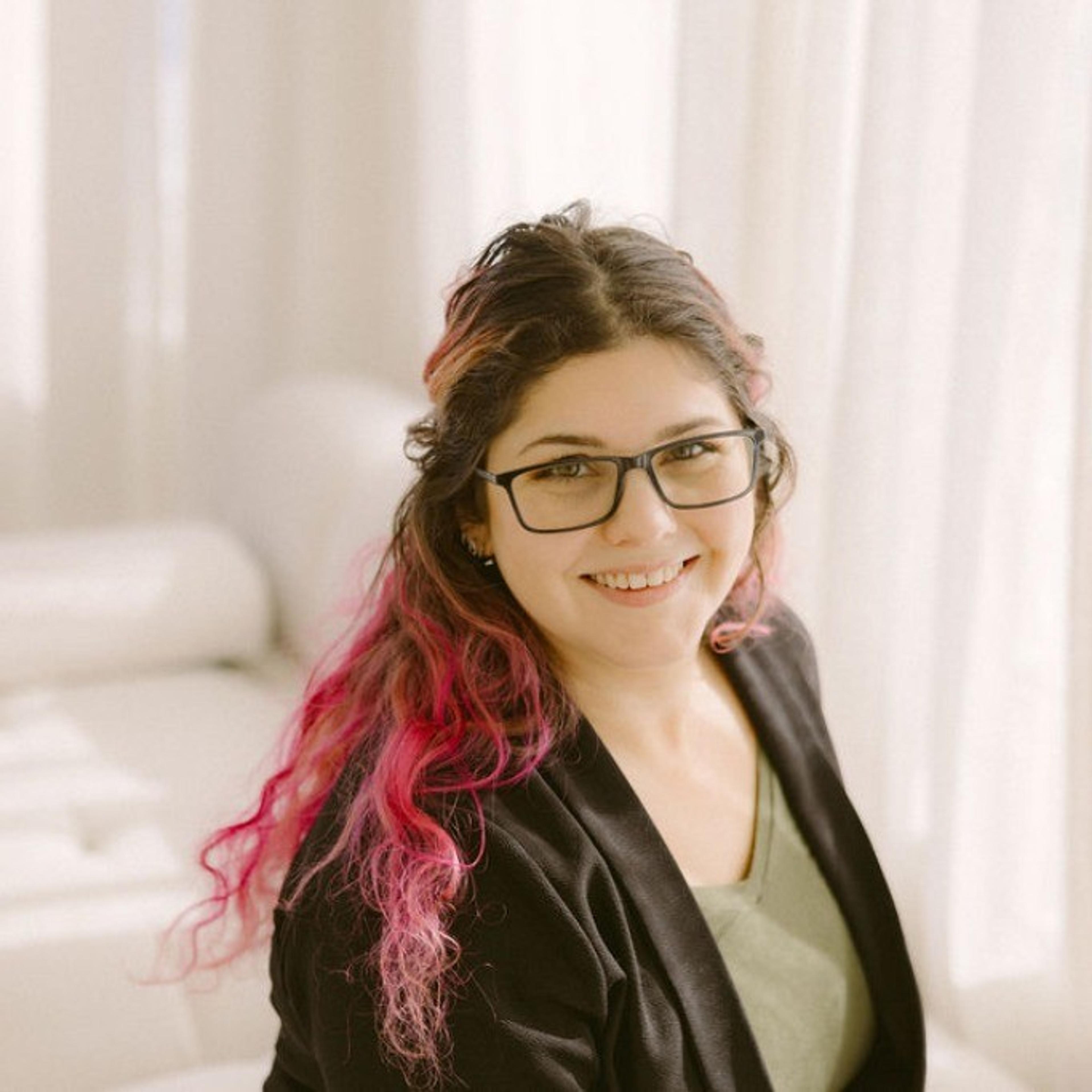 A young woman with glasses and pink-tinted hair smiles while sitting in a bright, airy room. She wears a black blazer over a green top.