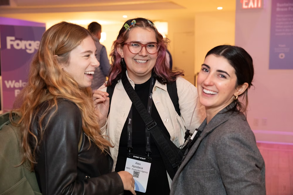 Tres mujeres sonríen y conversan en un evento. Una de ellas lleva una mochila y otra tiene una chaqueta gris.